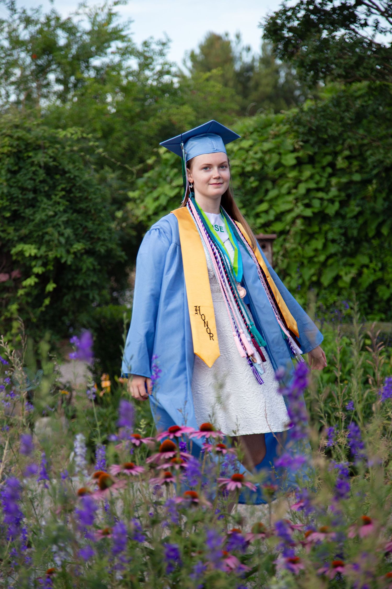 A smiling female graduate in blue cap and gown with honor stoles stands among colorful wildflowers in a lush garden.