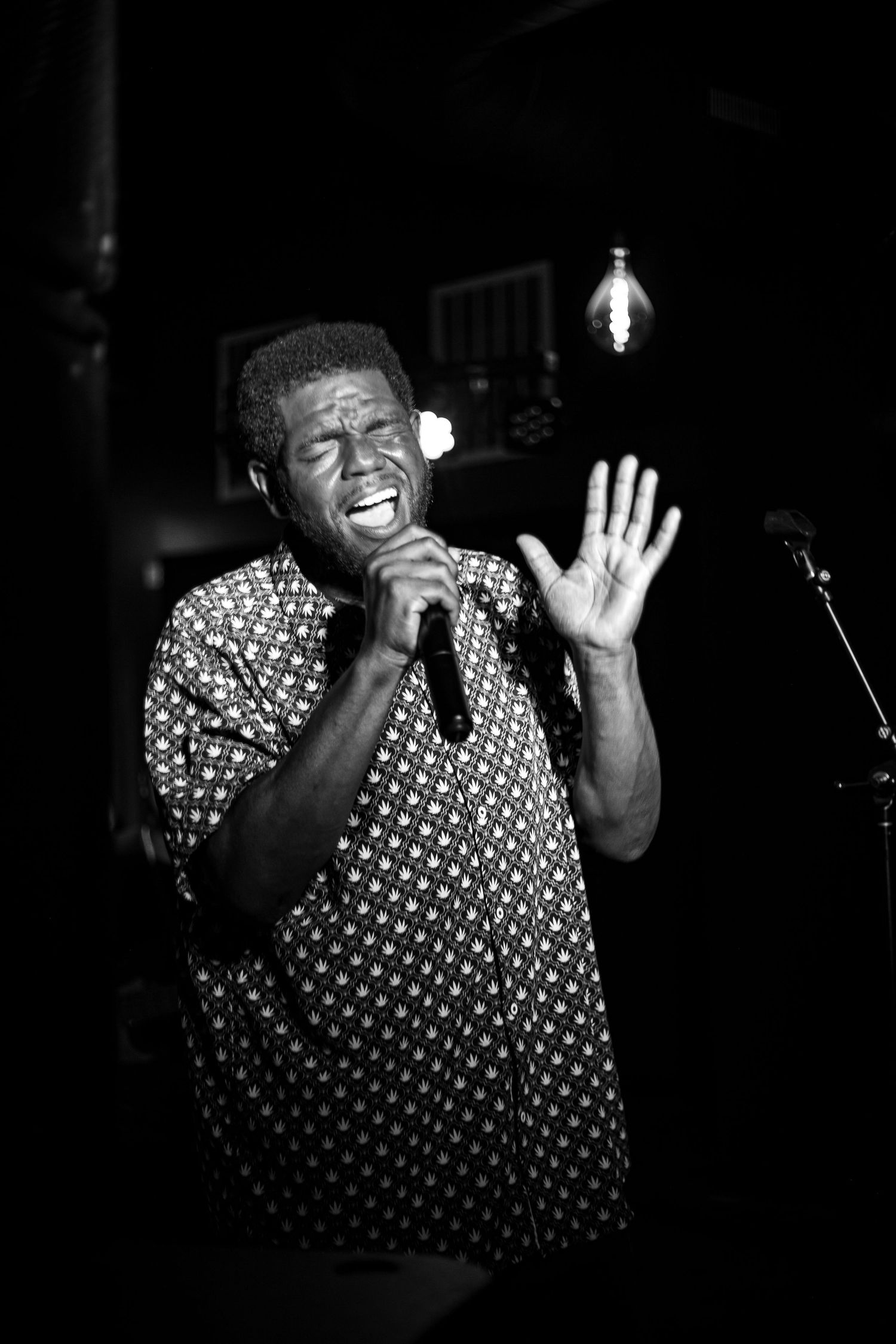 Black and white photo of a man singing passionately into a microphone on stage, hand raised.