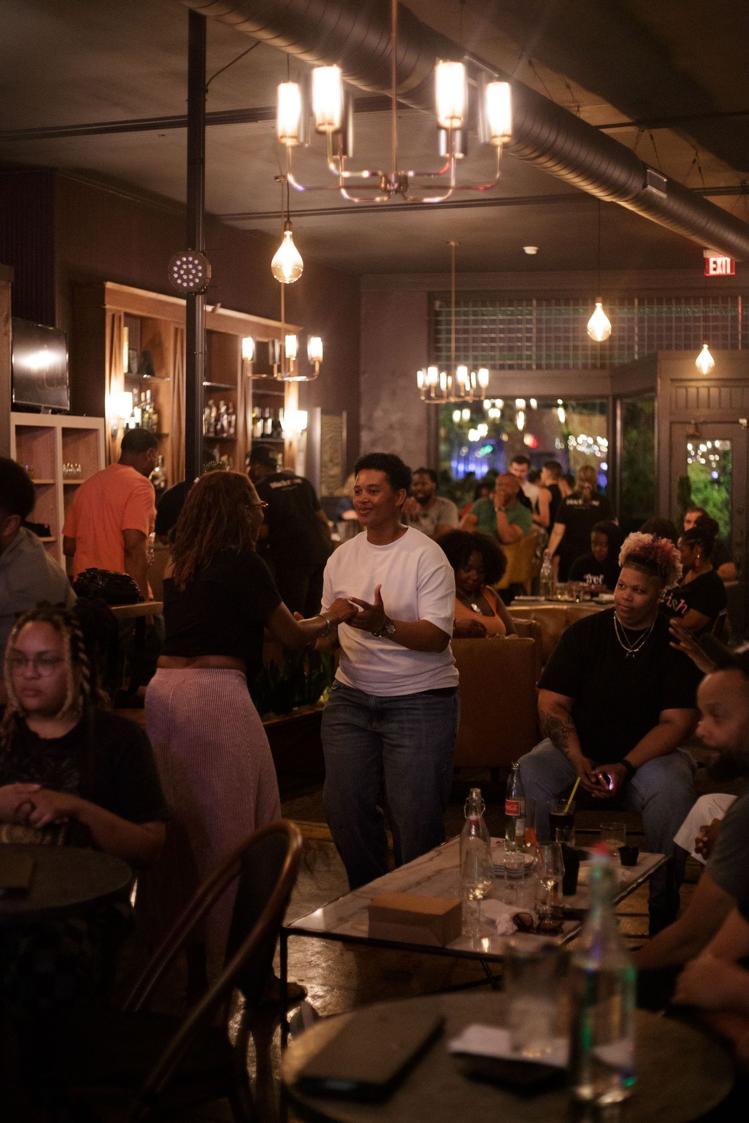 Busy restaurant interior with warm lighting, diverse guests dining, and a person in white tee standing among tables.