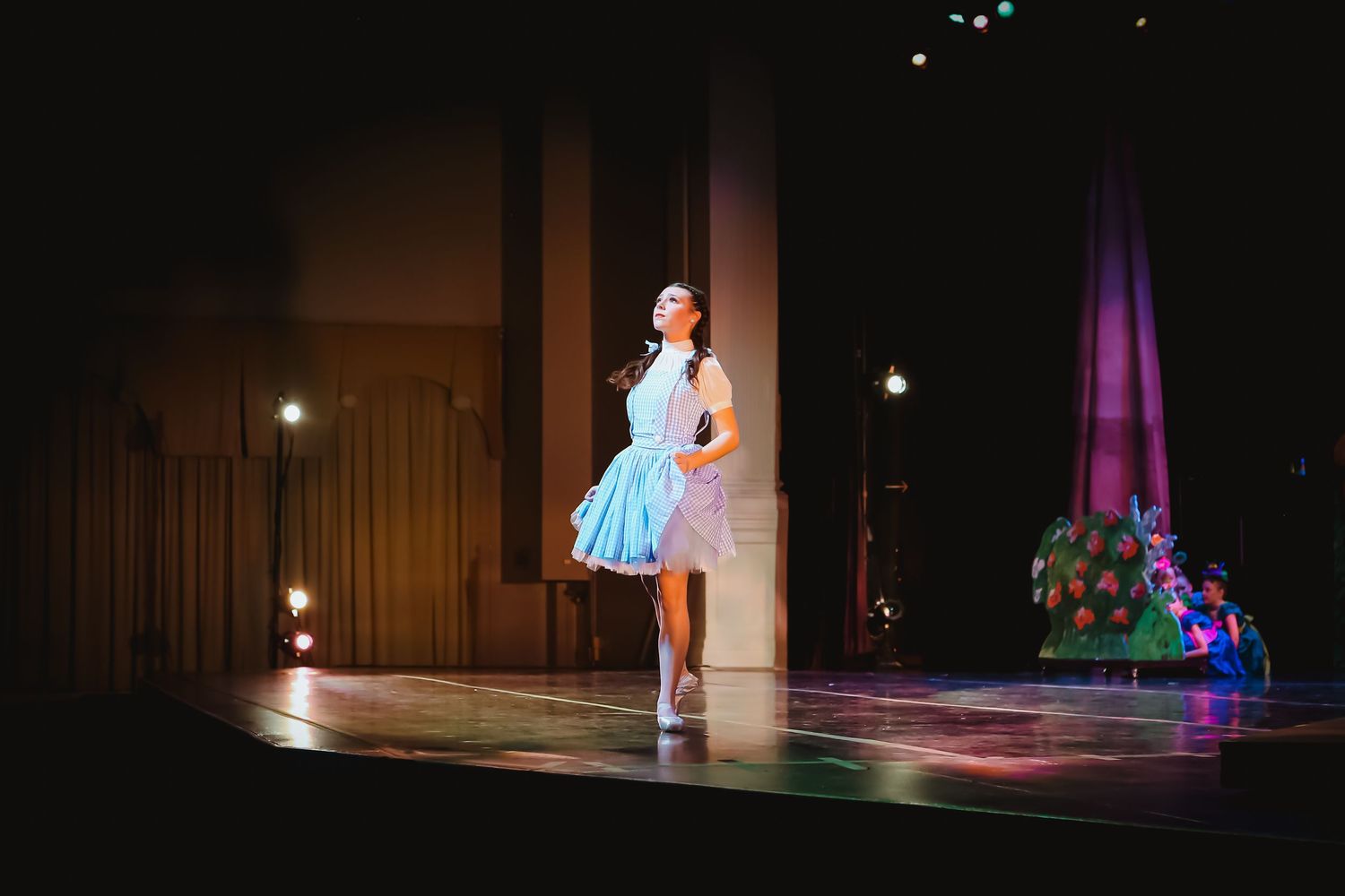 A performer in a blue dress sings on a dimly lit theater stage during a Wizard of Oz production.