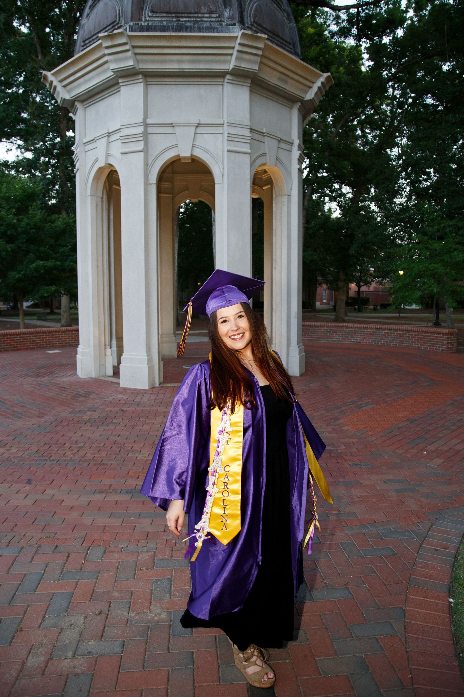 Young woman in purple graduation cap and gown smiling in front of a white gazebo on a brick campus plaza.