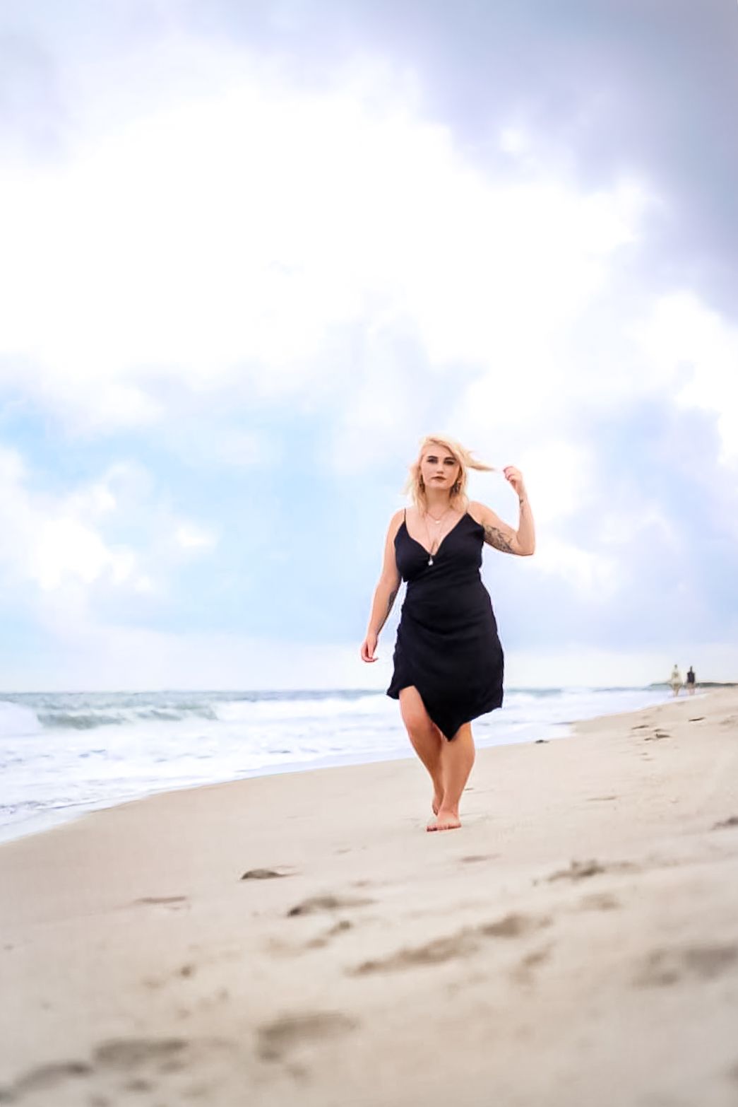 Blonde woman in black dress walking barefoot on sandy beach with ocean waves and cloudy sky behind her.