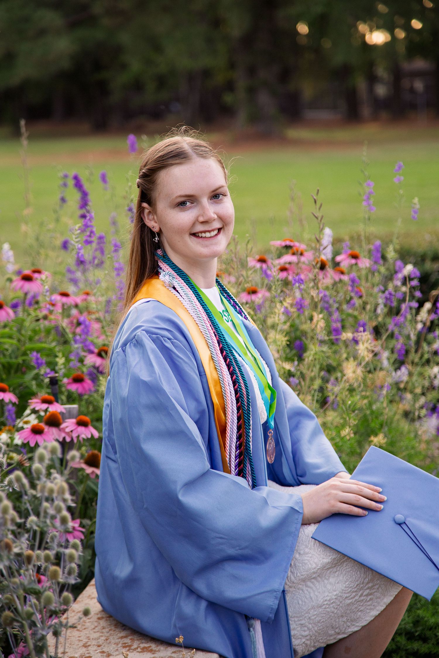 Smiling graduate in blue cap and gown with colorful stoles sits among purple and pink flowers outdoors.