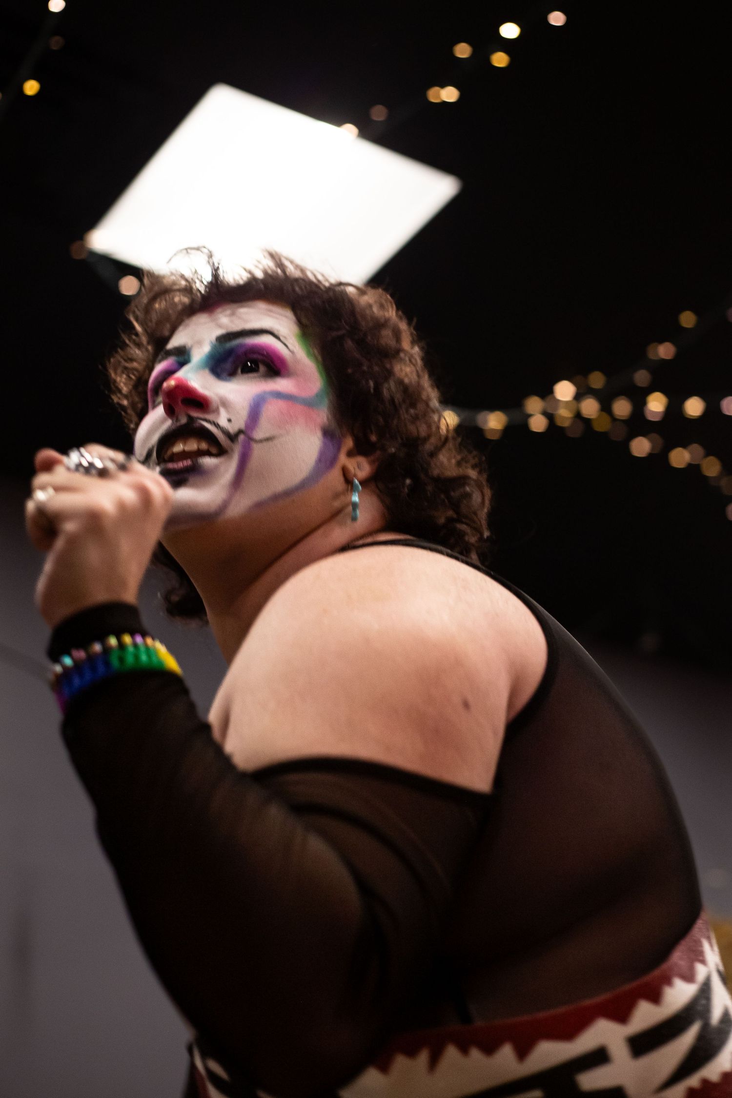 Performer with colorful clown face paint singing into microphone on dark stage with bright light backdrop.