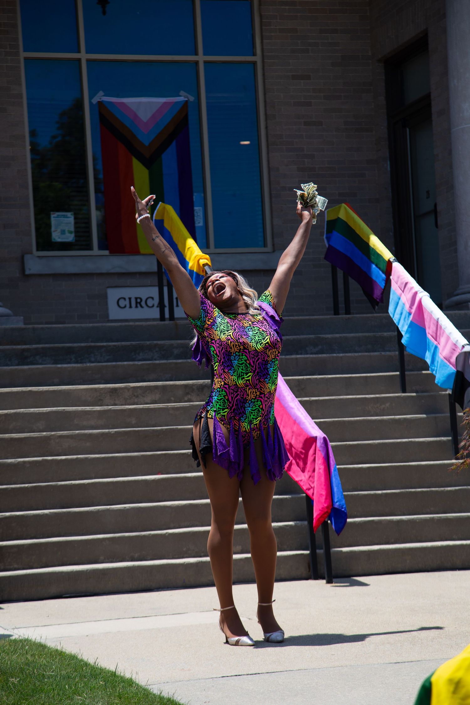 Person in colorful sequin dress celebrates at Pride event with rainbow and transgender flags on building steps.