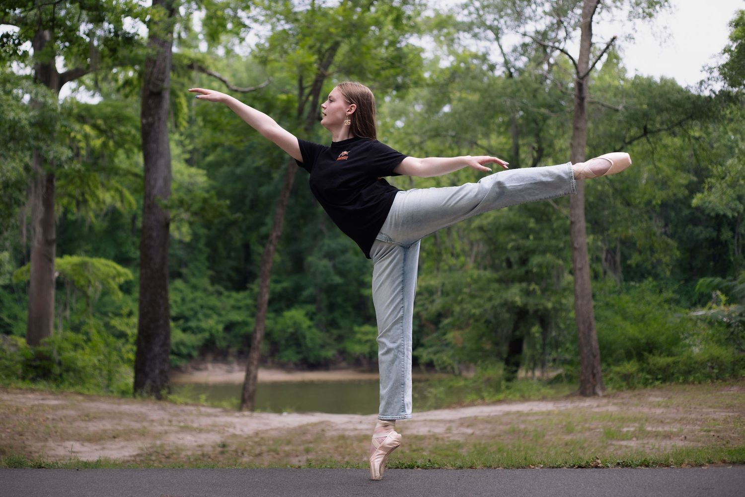 A ballet dancer performs an arabesque on pointe outdoors, wearing black top and gray pants, surrounded by lush green trees.
