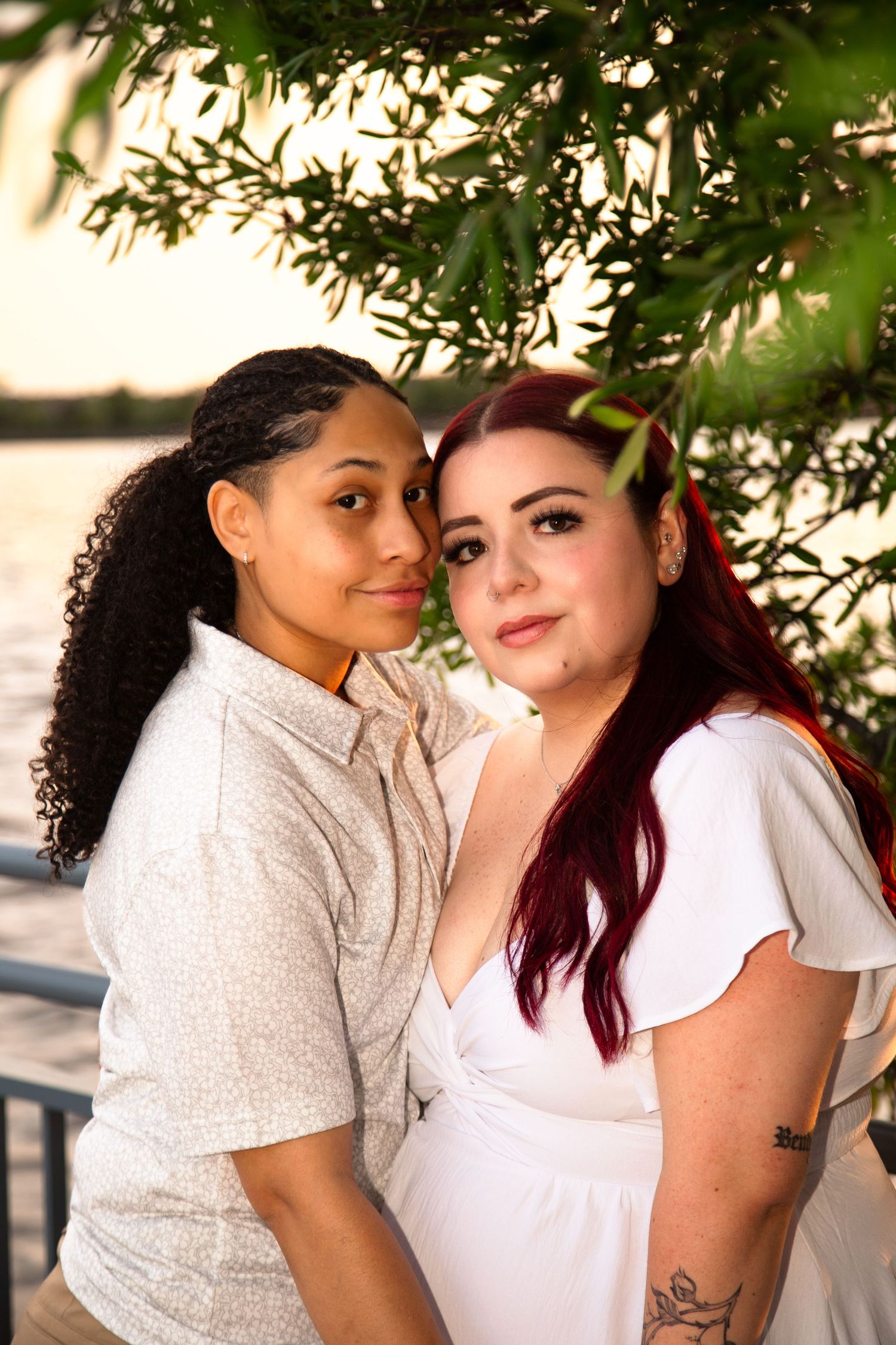 Two women posing together under a willow tree near a waterfront at sunset, one in white dress with red hair.