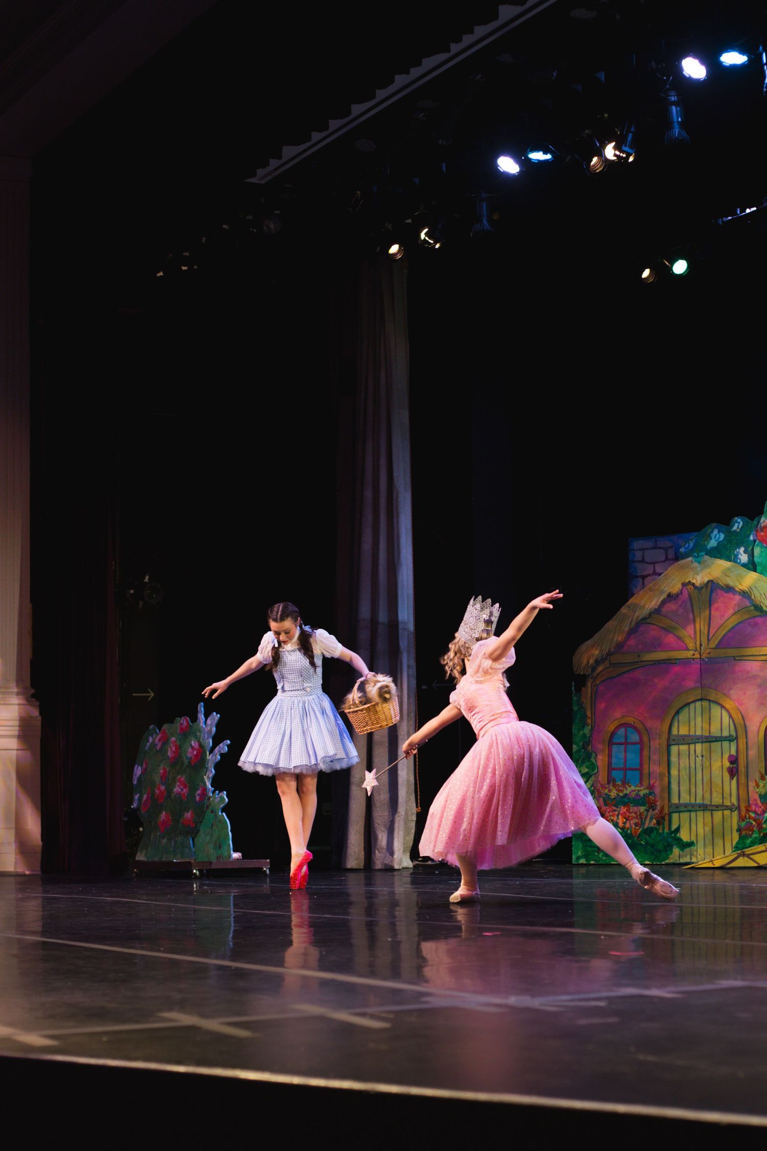 Two dancers perform Wizard of Oz ballet on stage, one as Dorothy in blue dress with basket, other as Glinda in pink gown.