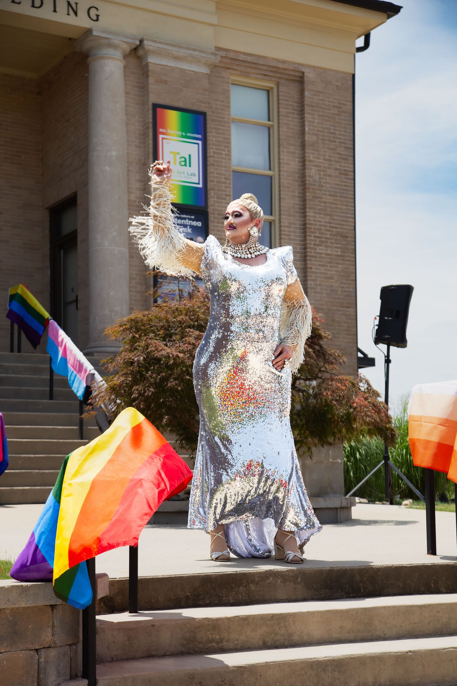 Drag queen in sequined gown raises fist at Pride event with rainbow flags on sunny building steps.