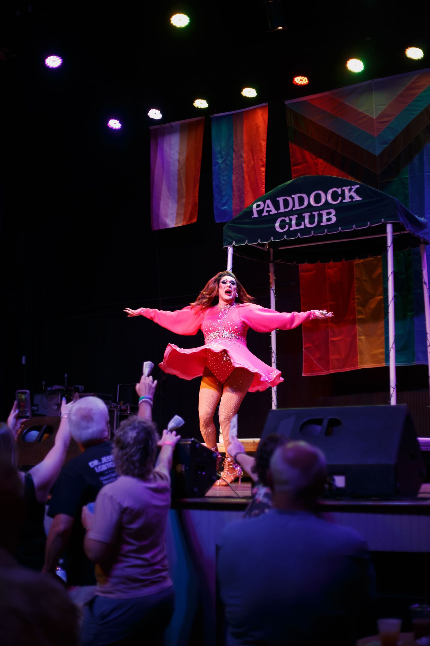 Drag performer in pink costume dancing on stage at Paddock Club with rainbow Pride flags backdrop.