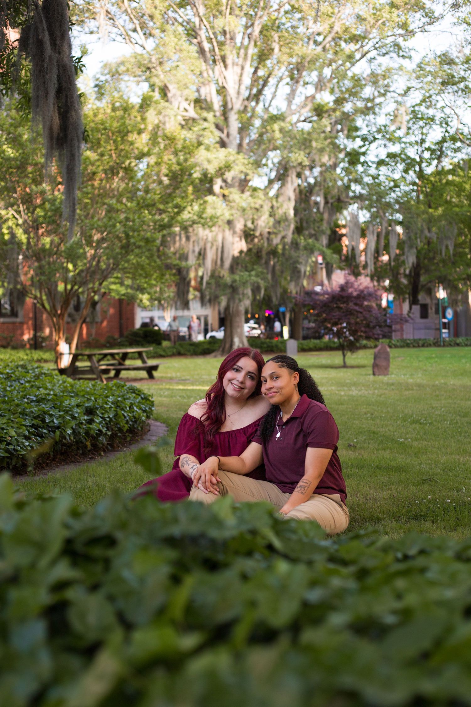 Two young women sitting together on green grass in a sunlit park with Spanish moss-draped trees.