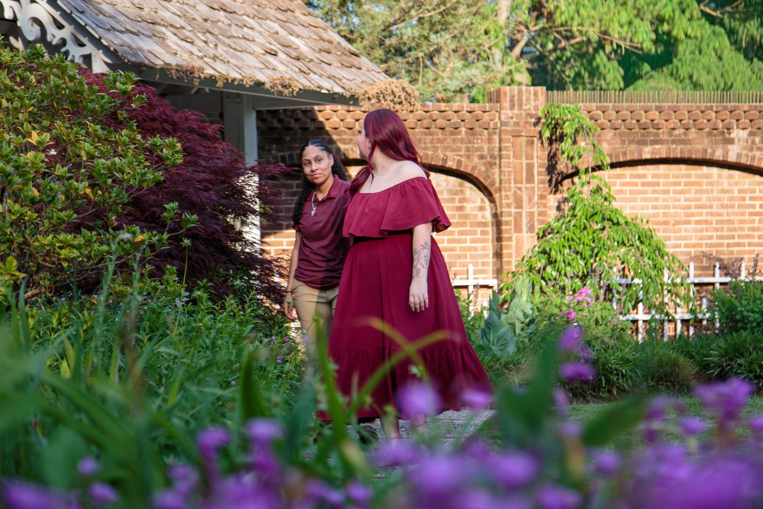 Two women in burgundy dresses standing in colorful garden near brick wall.