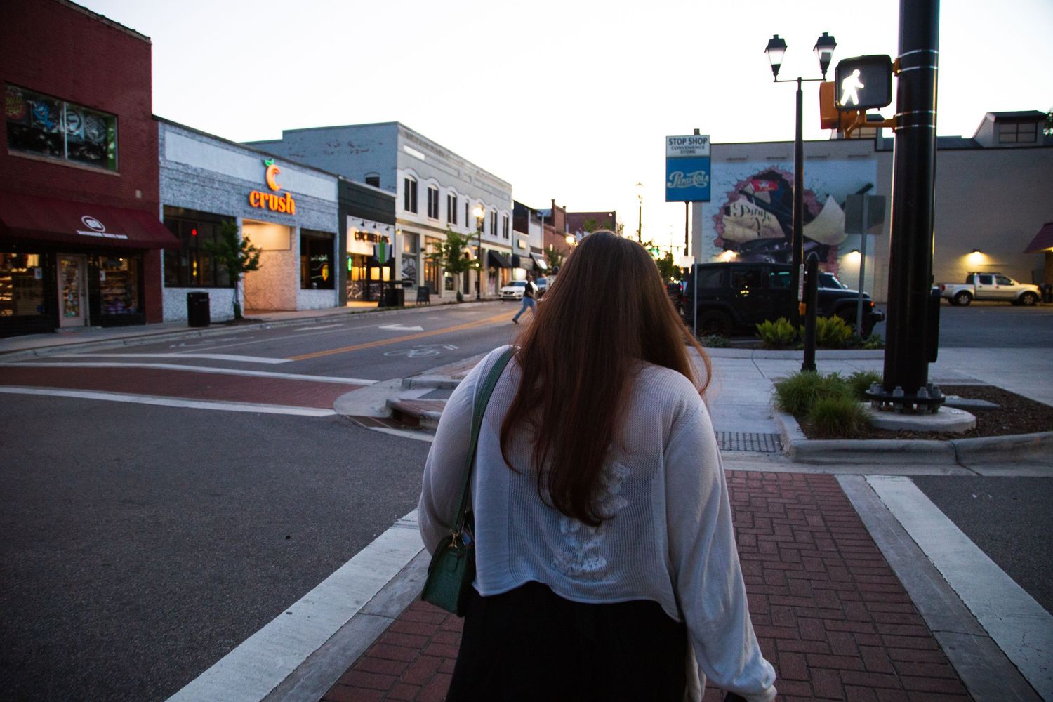 Woman with long hair walking along a quiet downtown street at dusk, passing storefronts and a street lamp.