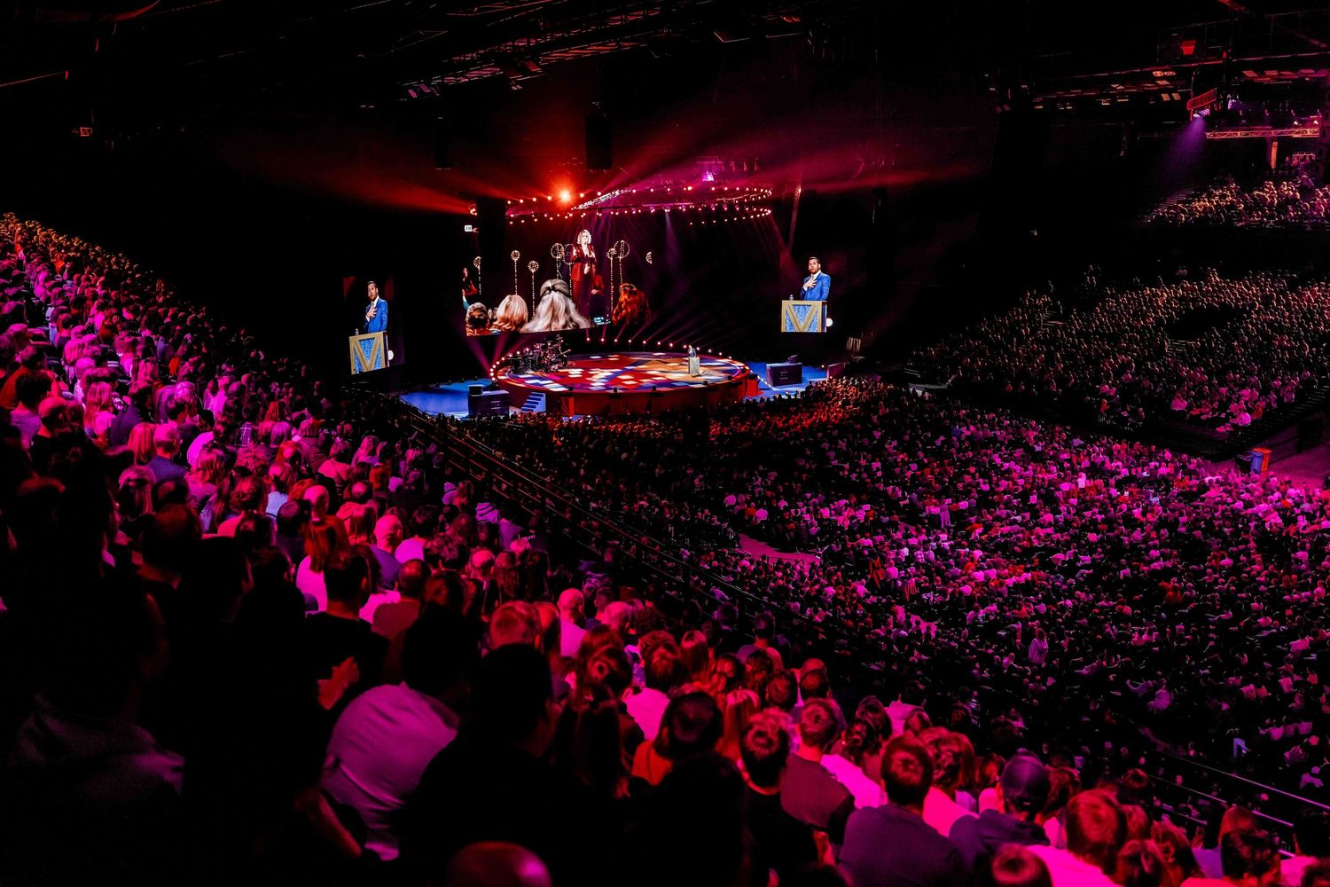 Large arena concert with packed crowd bathed in pink and purple lights, stage glowing at center.