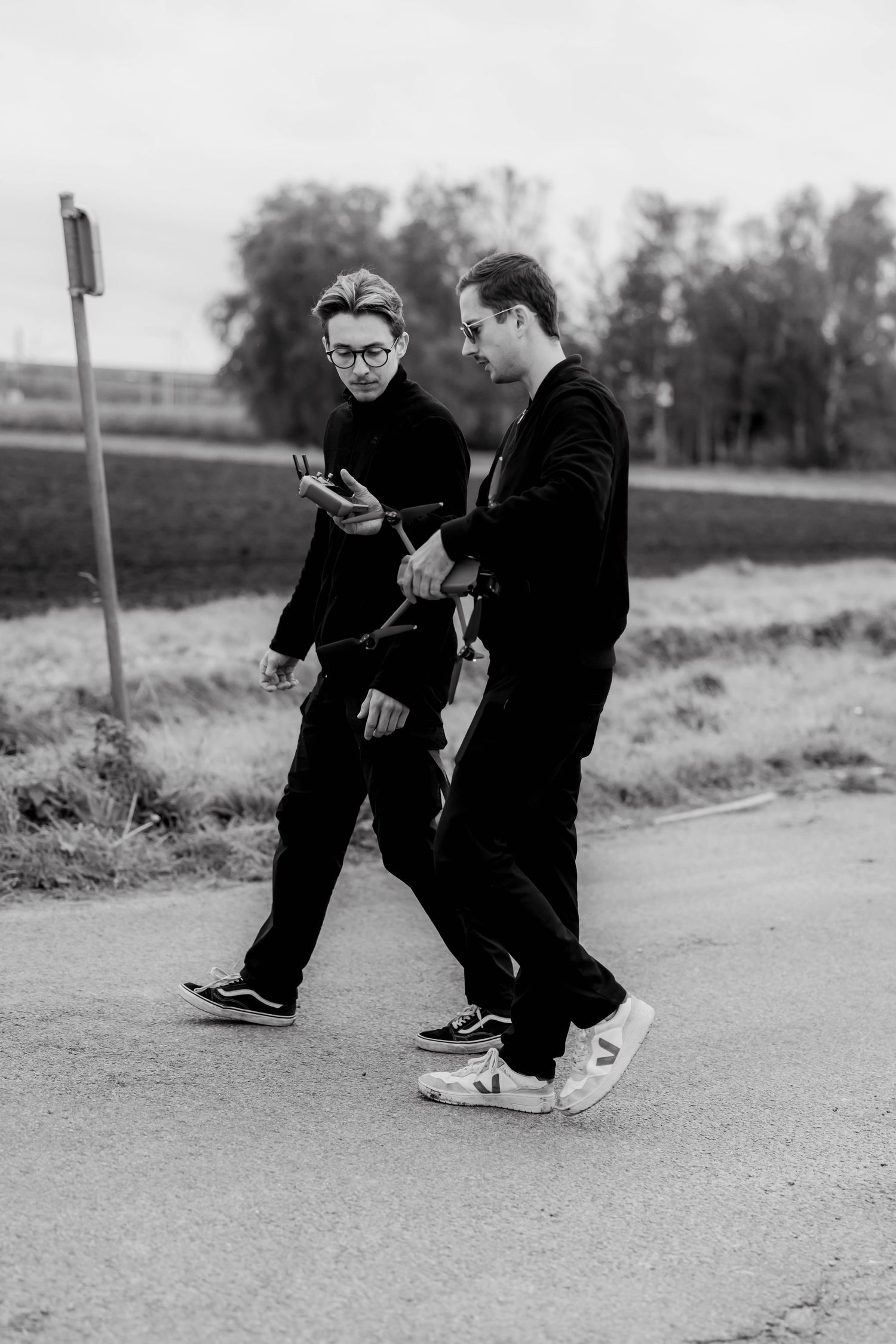 Two young men dressed in black walk together on a road, one checking a smartphone, in a black and white photo.