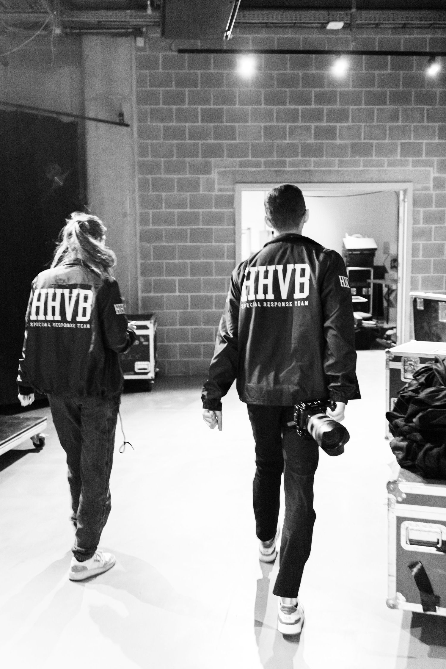 Two people wearing HHVB jackets walk backstage in a black and white photo, one carrying a camera.