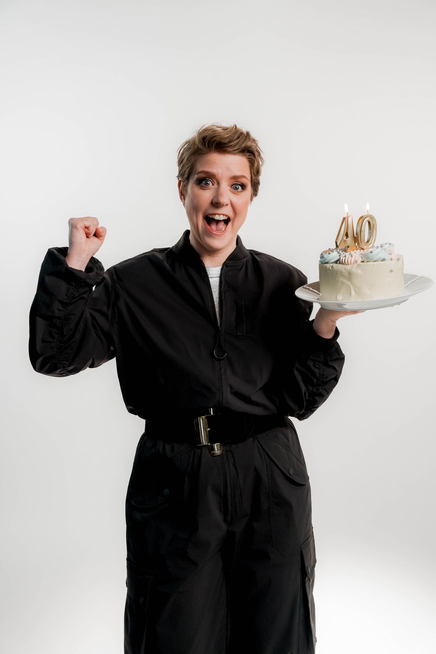 Excited woman in black outfit holding a 40th birthday cake, raising her fist in celebration on white background.