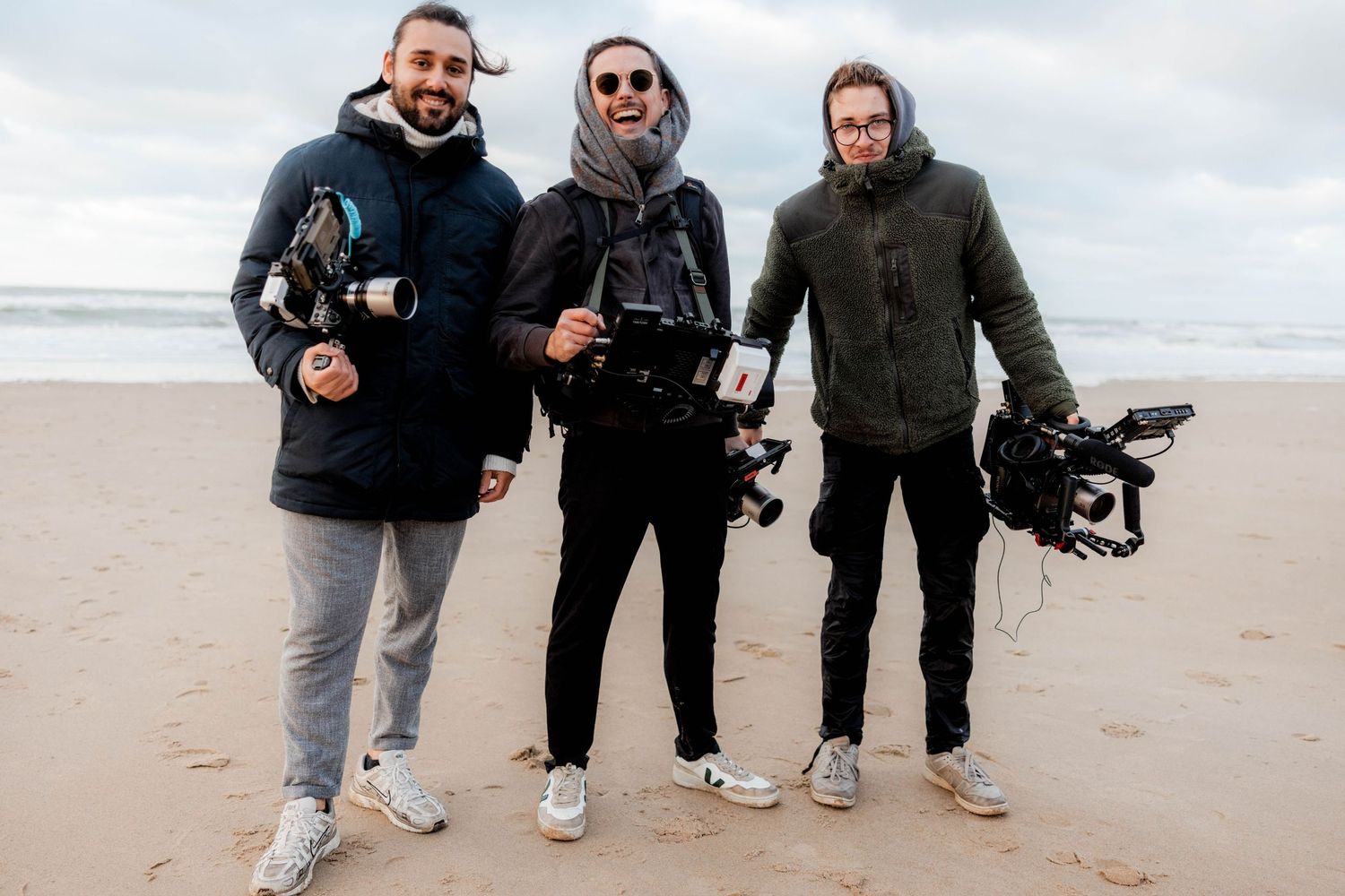 Three men holding professional cameras pose together on a sandy beach with an overcast sky.