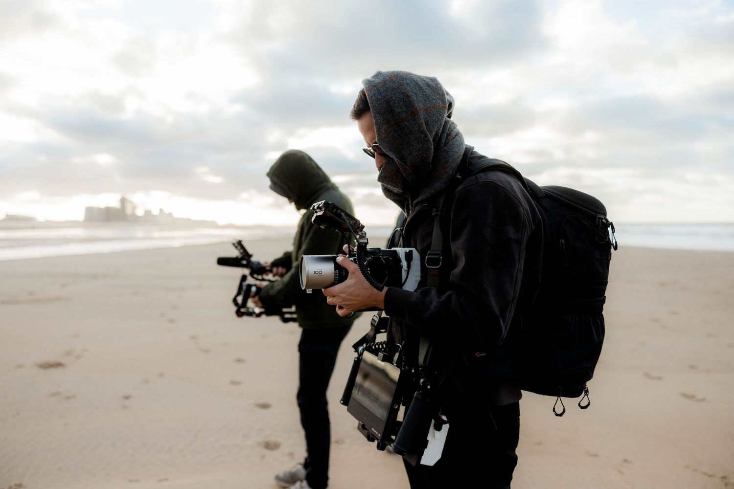 Two videographers in black hoodies filming on a beach with professional cameras on a cloudy day.