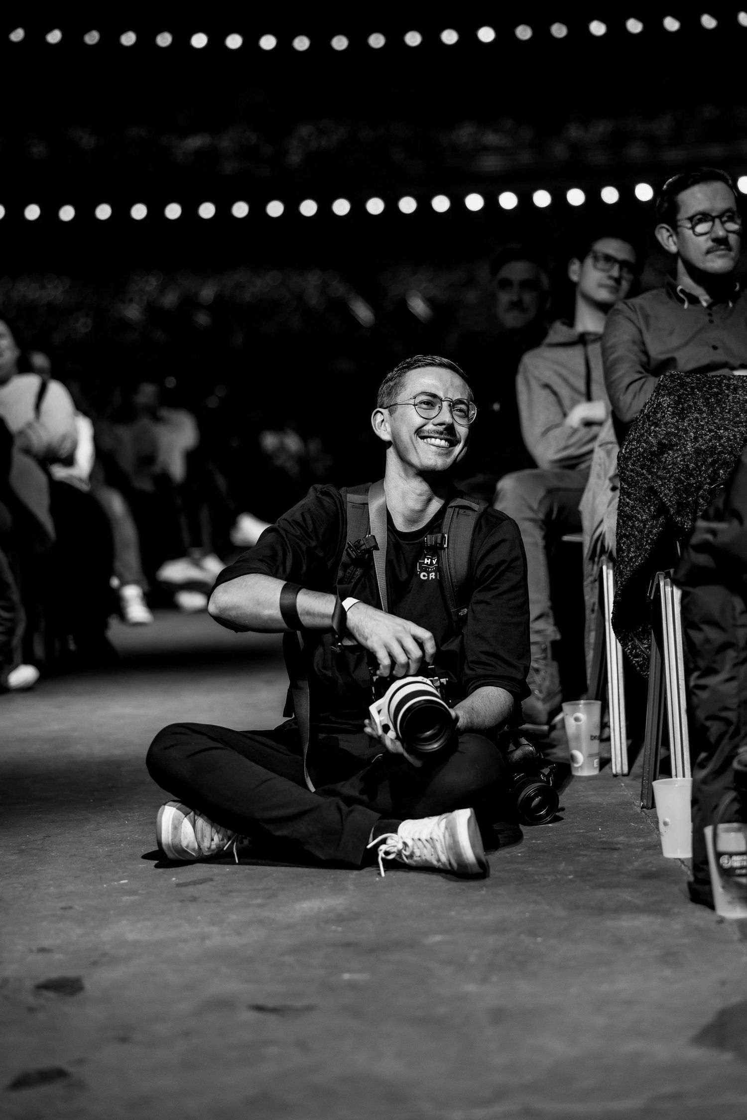 Smiling young man sitting cross-legged on floor holding camera at event, black and white photo.