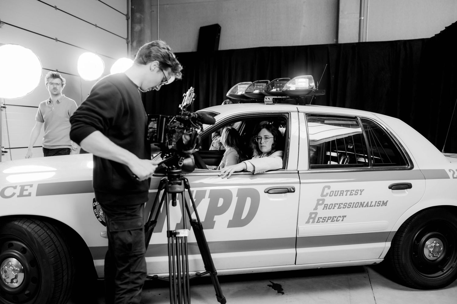 A cameraman films a woman sitting in an NYPD police car on a professional film set in black and white.