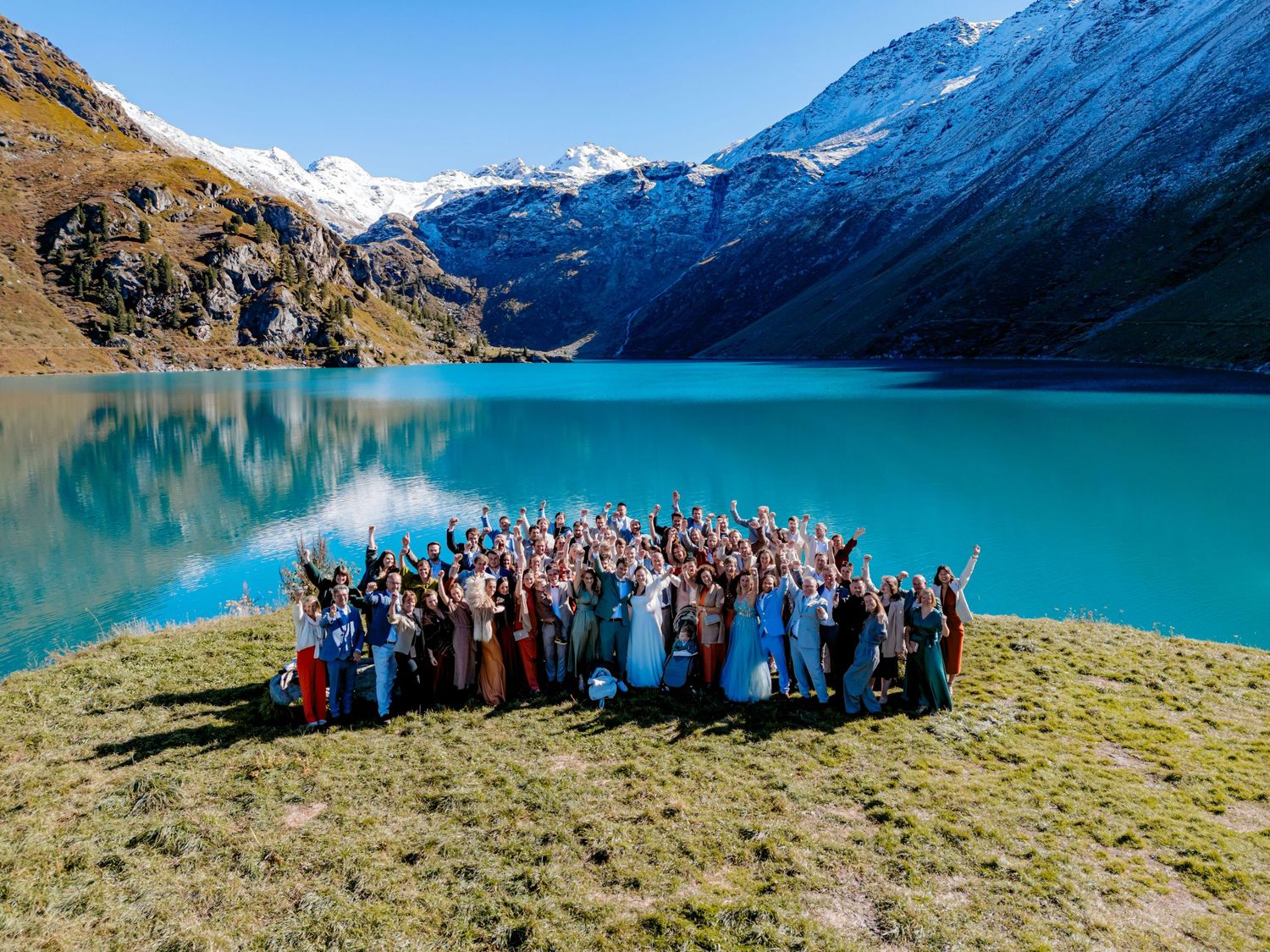 Aerial view of a large wedding group celebrating by a turquoise alpine lake surrounded by snow-capped mountains.