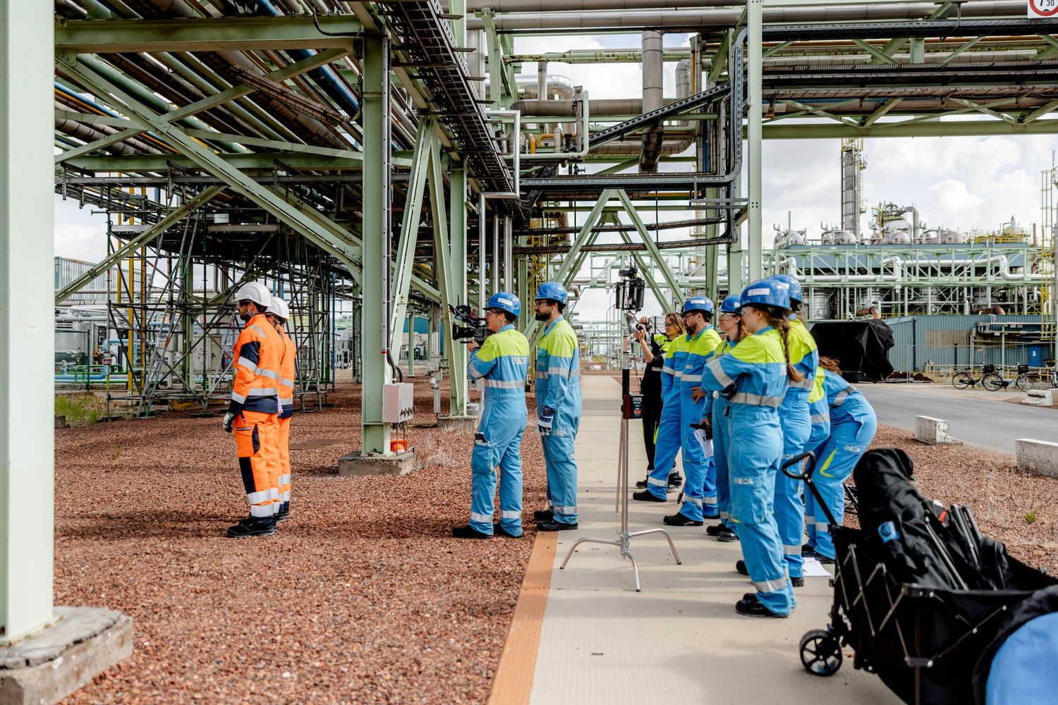 Industrial plant workers in blue safety gear receiving a briefing outdoors at a chemical refinery facility.