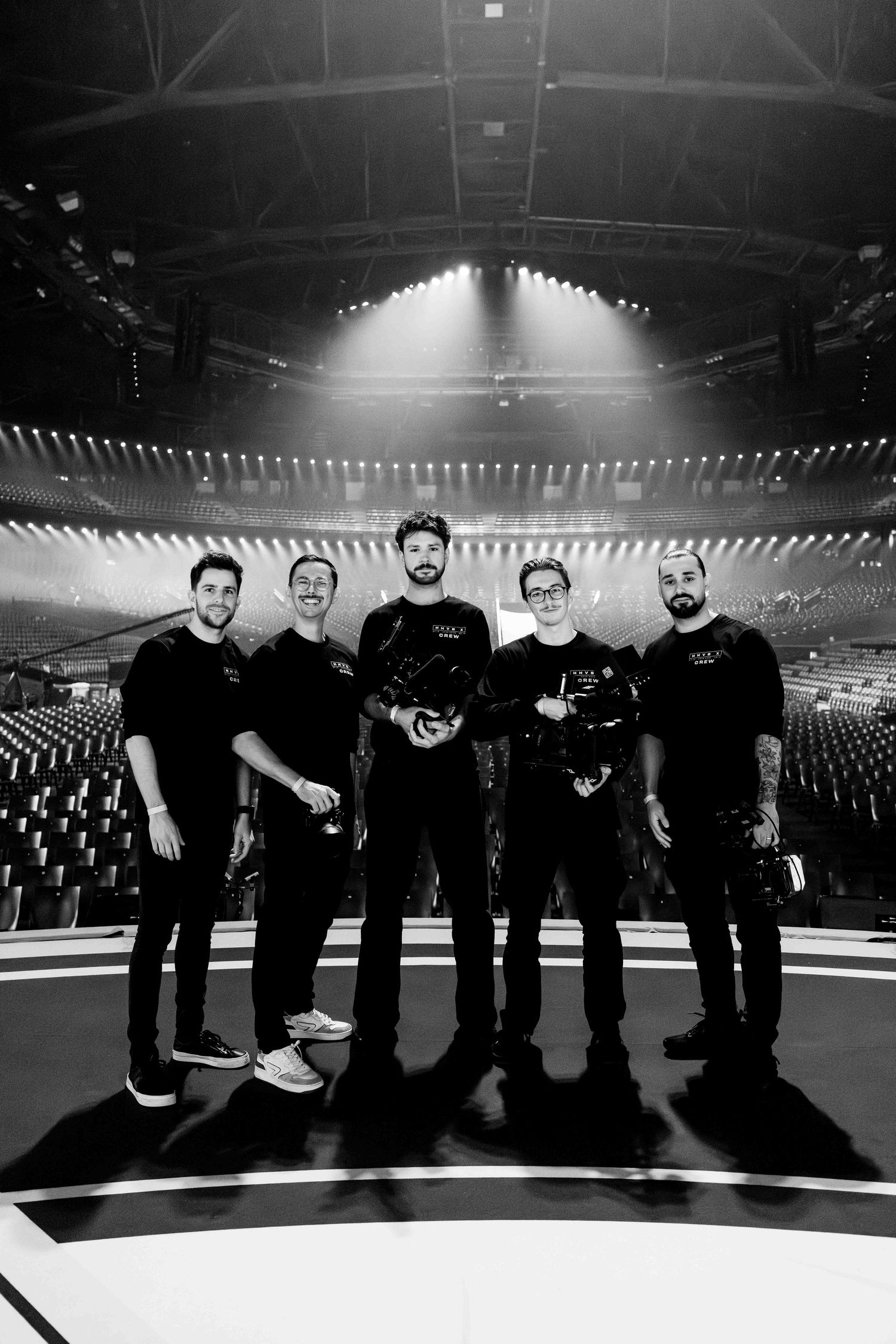 Five people in black attire pose together on a stage in a large arena, captured in black and white.
