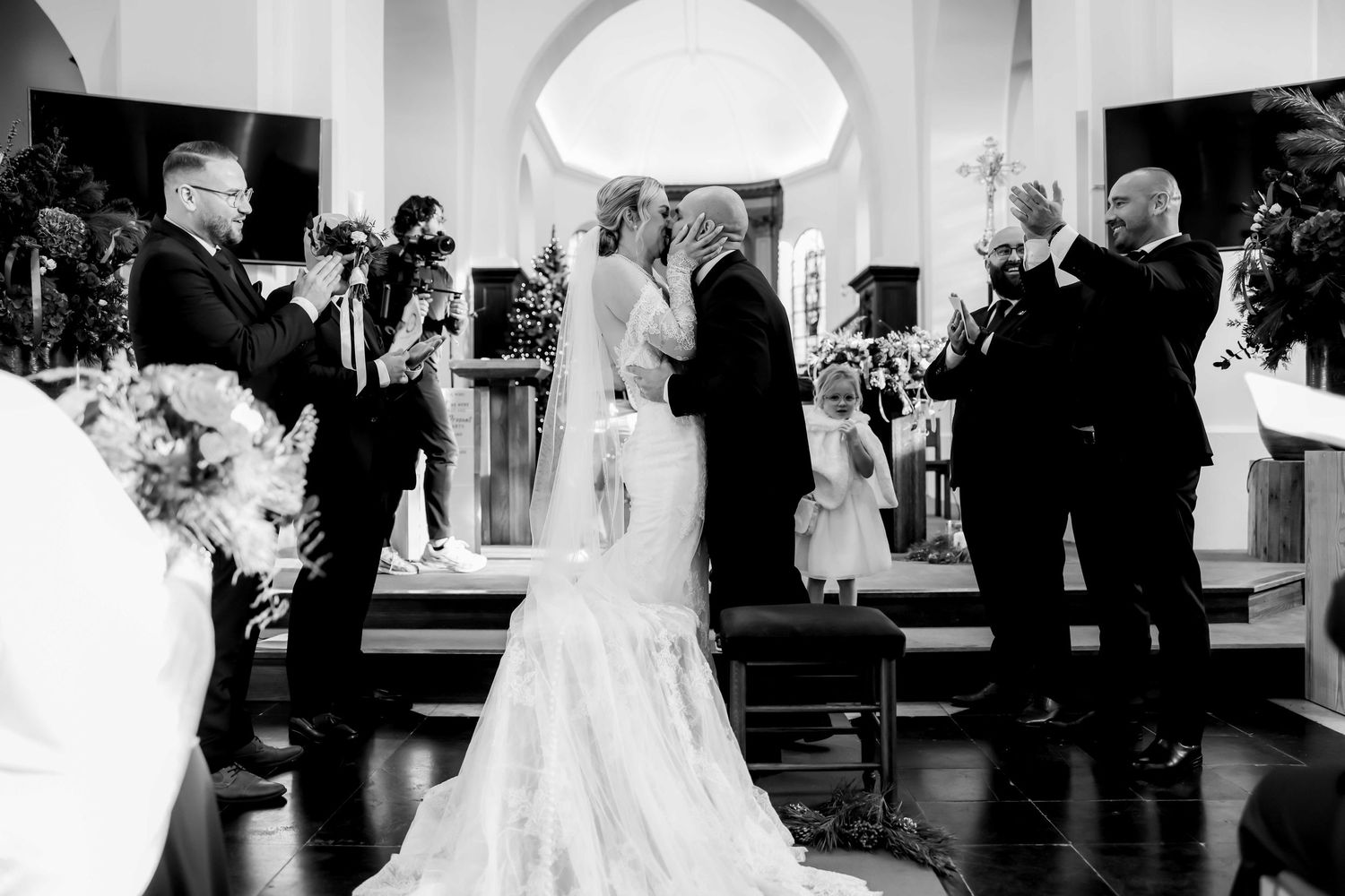 Black and white wedding ceremony photo of bride and groom at church altar with guests and flower girl.