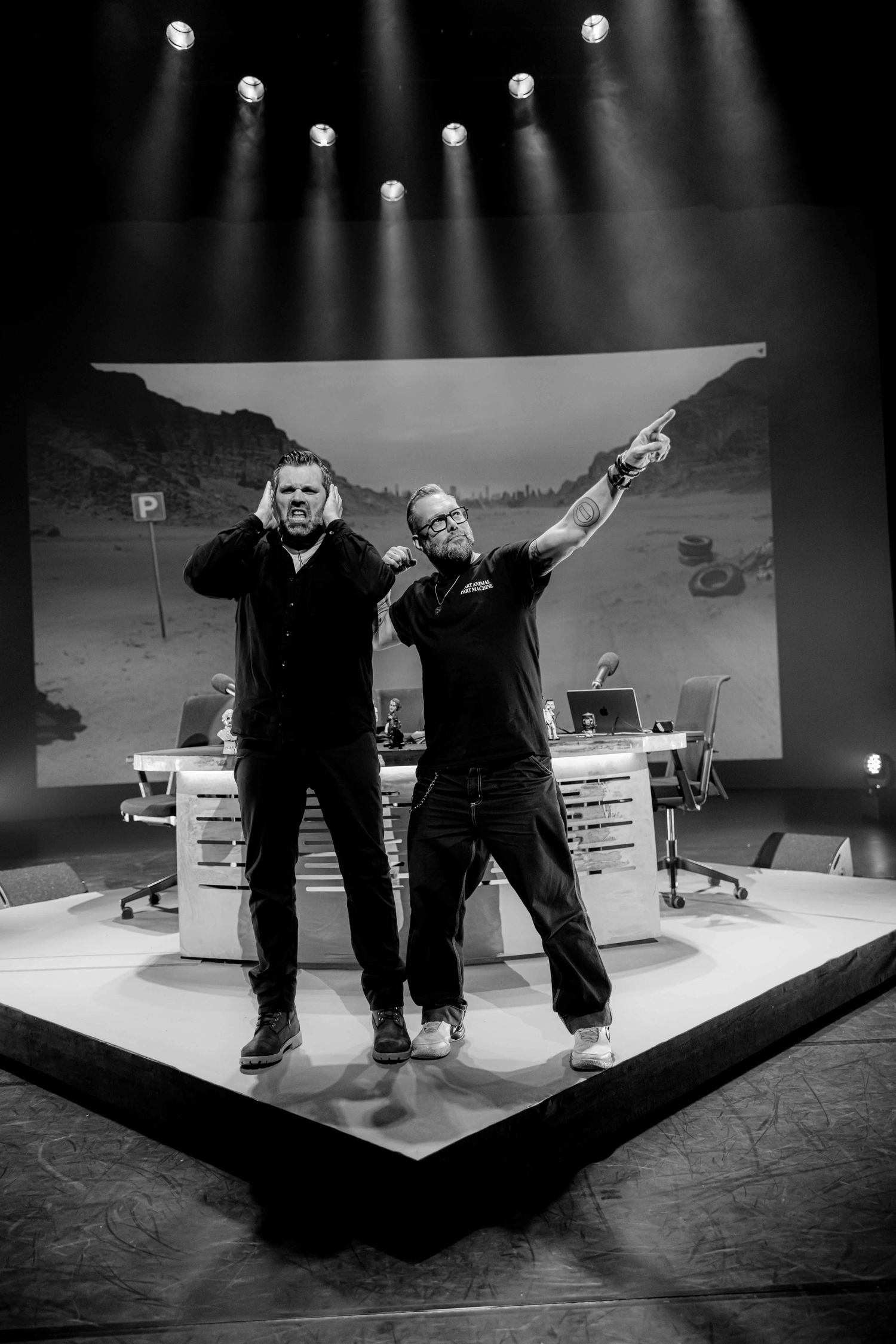Two men pose dramatically on a stage with studio lighting and a desert backdrop in a black and white photo.