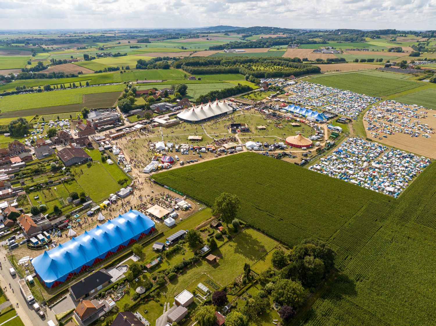 Aerial view of a large outdoor festival with blue tents, camping areas, and surrounding green farmland.