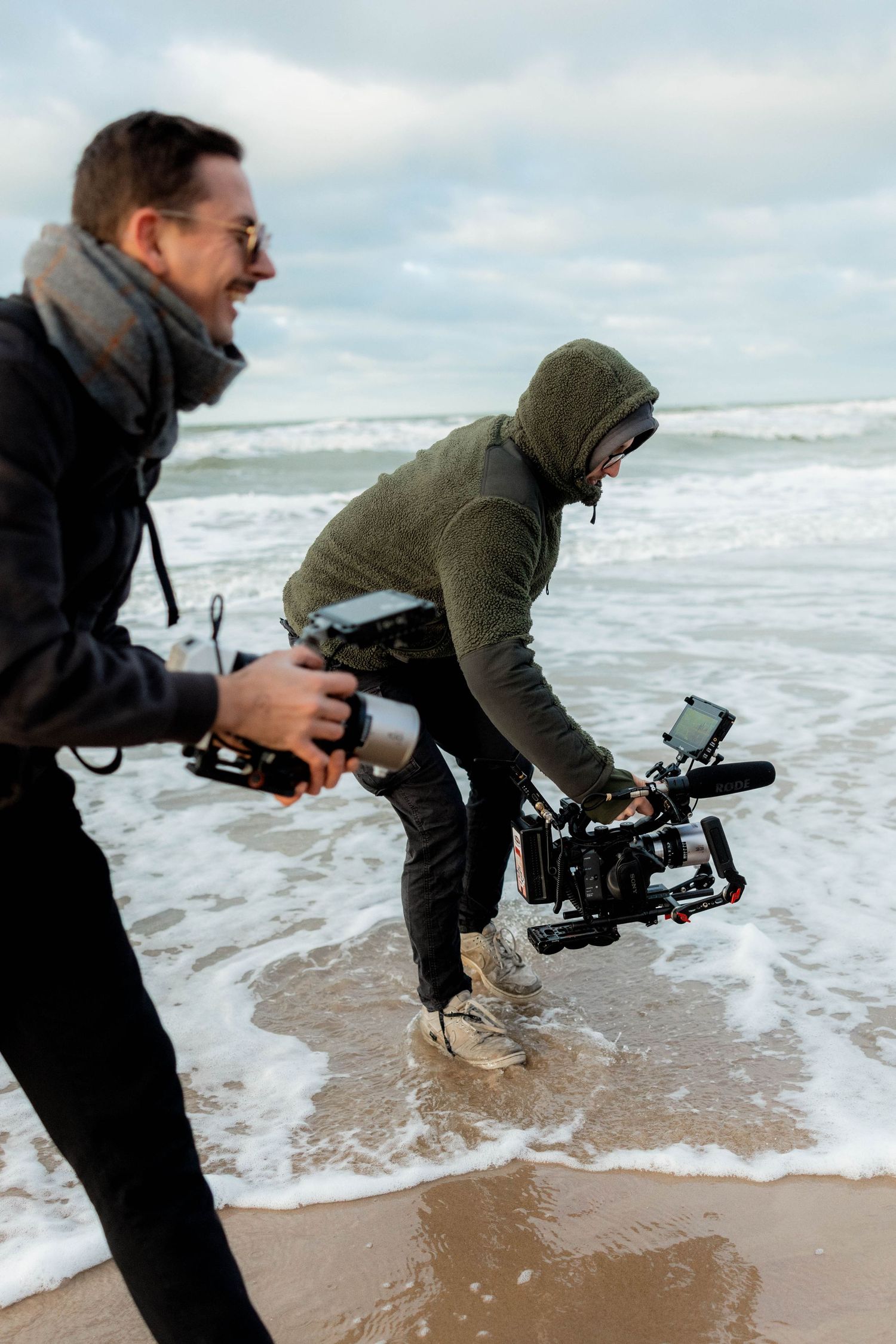 Two videographers filming on a beach with professional camera rigs, waves washing ashore on a cloudy day.