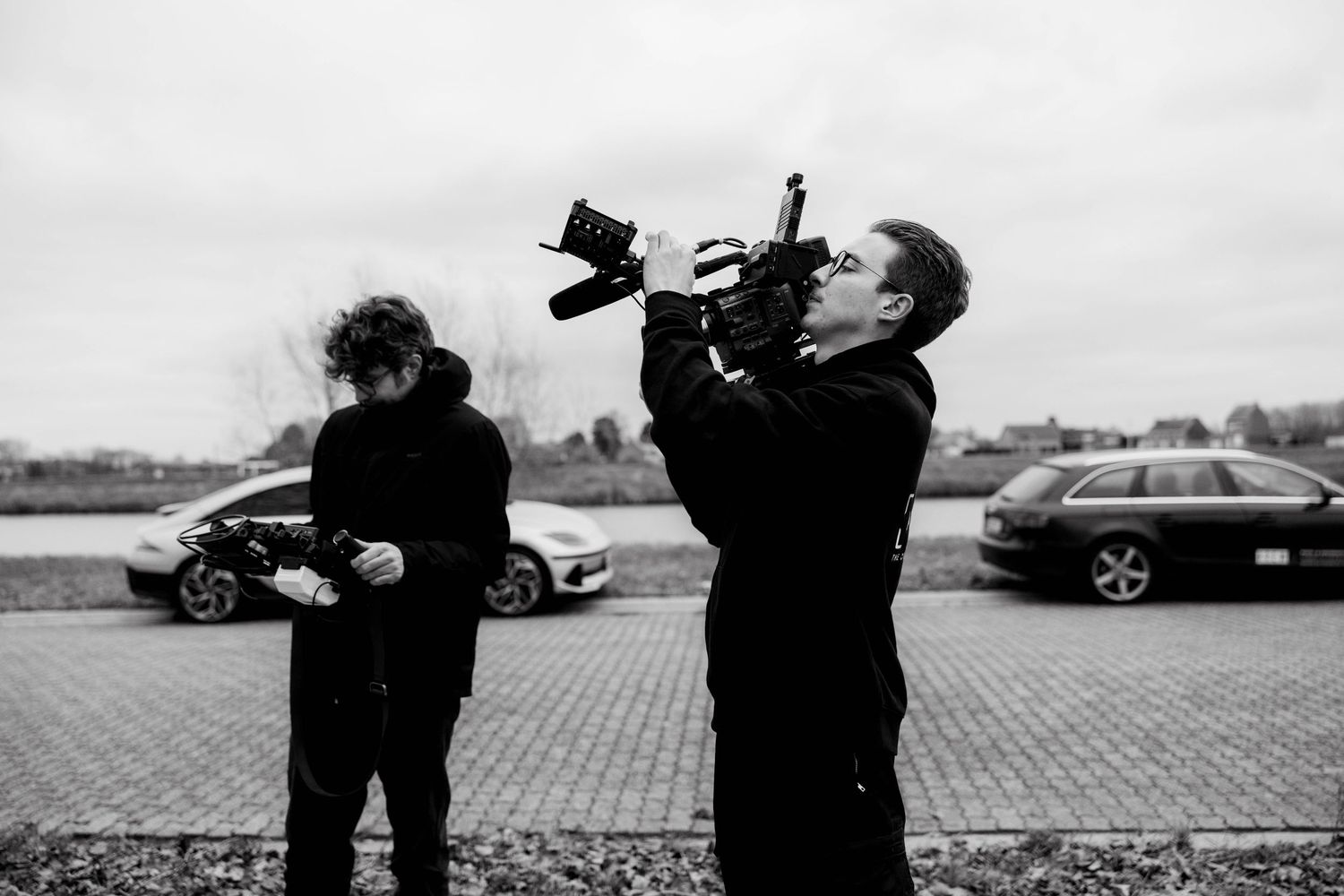 Two filmmakers in black clothing operate a professional camera outdoors near parked cars on a cloudy day.