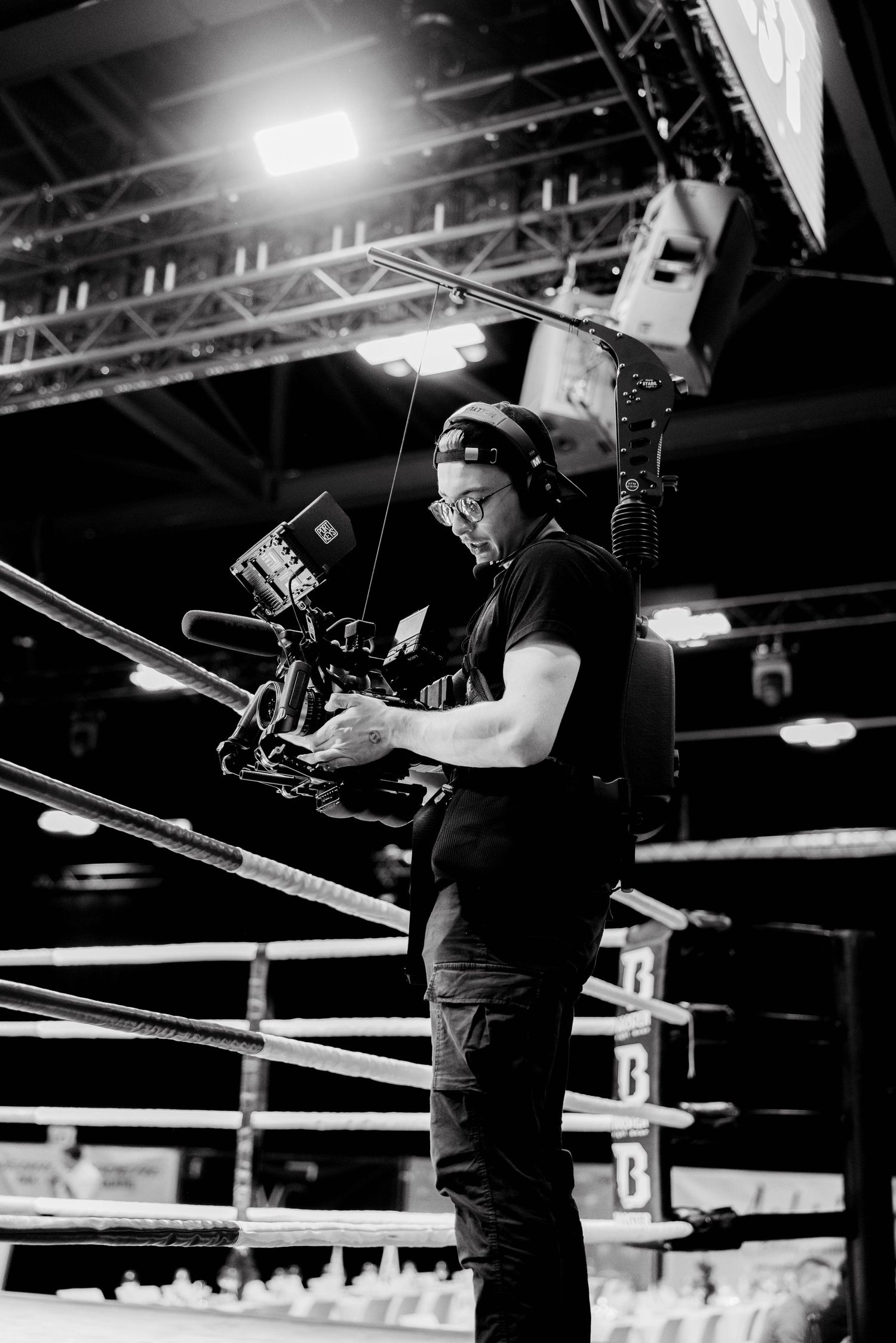 Black and white photo of a cameraman filming at a boxing ring event, wearing headphones and holding a professional video camera.