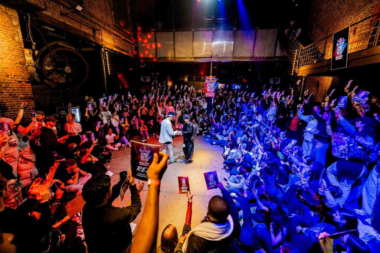 Energetic concert crowd fills a brick-walled venue as performers take the stage under colorful red and blue lights.
