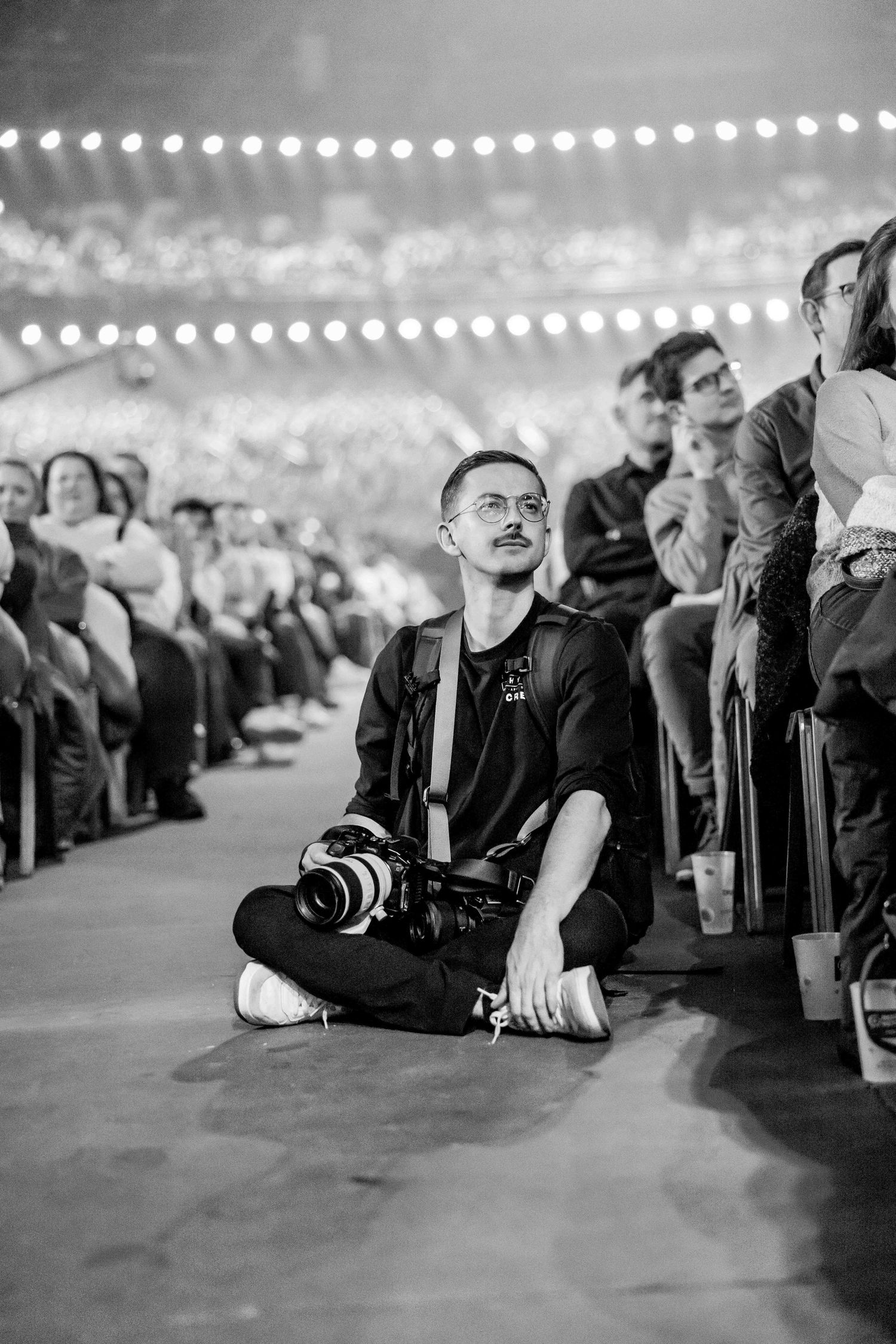 Black and white photo of a photographer sitting cross-legged on the floor at a crowded event, camera around neck.