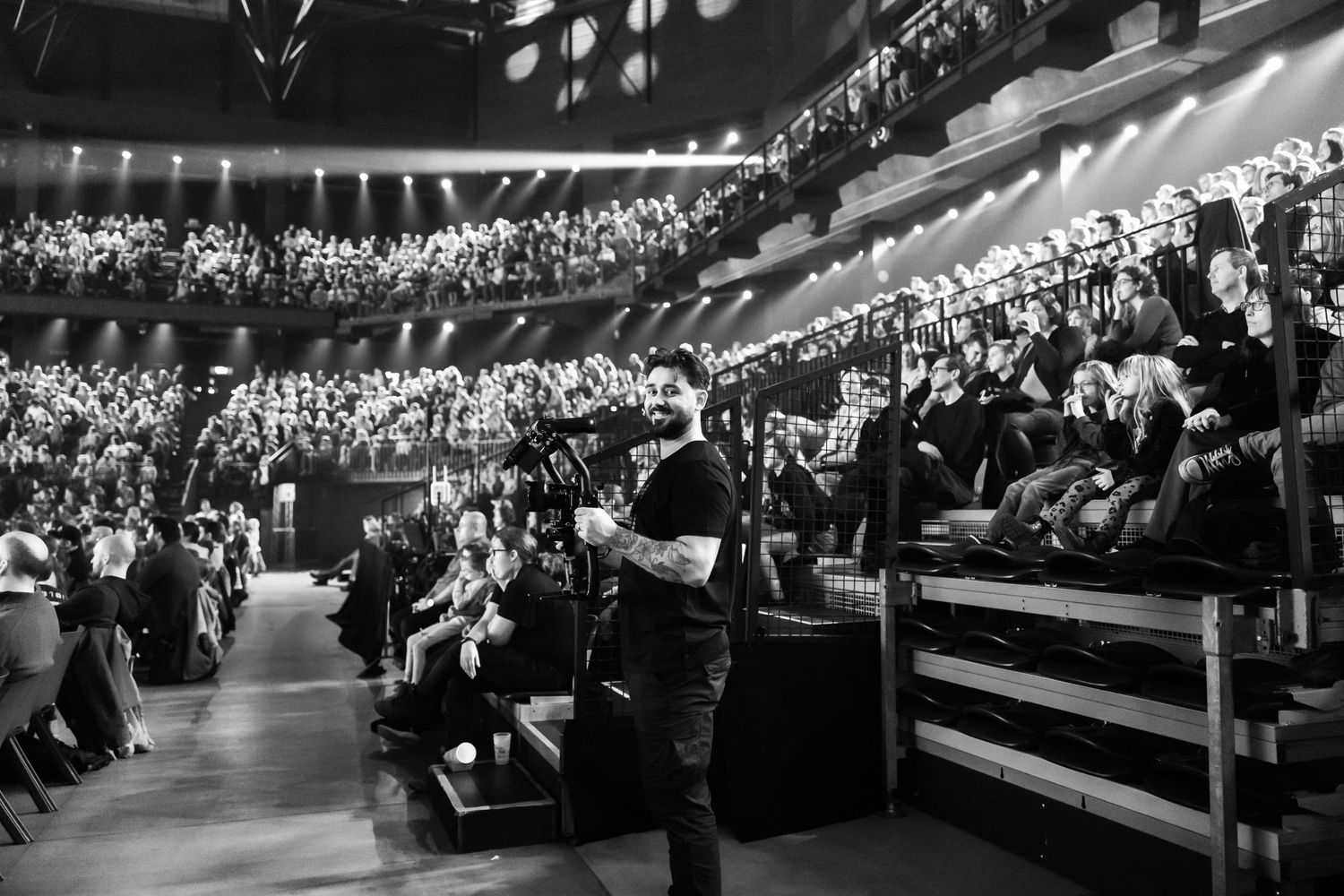 Black and white photo of a cameraman filming a packed concert hall audience from the stage floor.