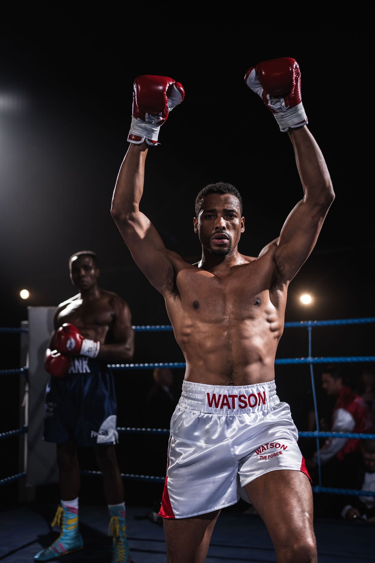 A boxer wearing Watson shorts raises his arms in victory inside a boxing ring.