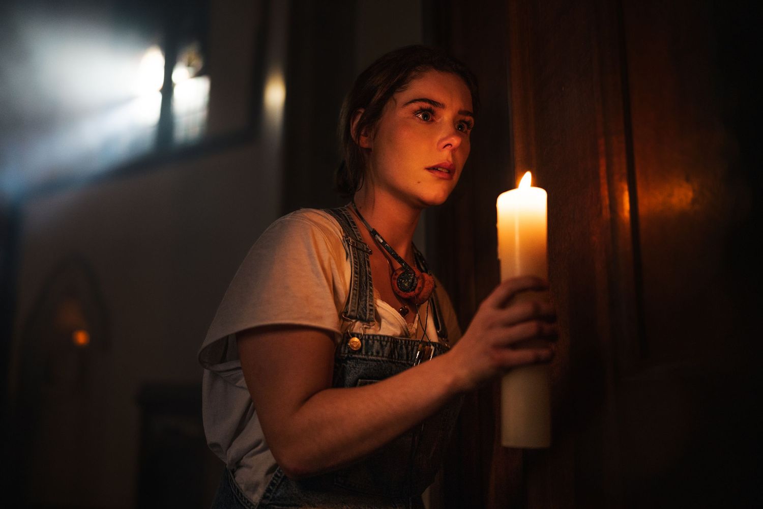 A young woman in overalls holds a large white candle in a dark, atmospheric setting.