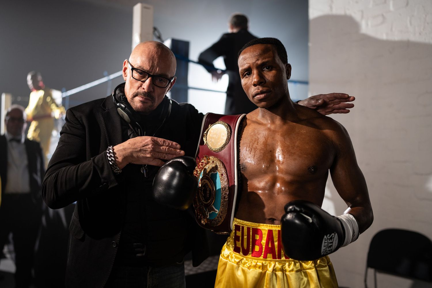 Boxer holding championship belt with trainer in locker room after victory.