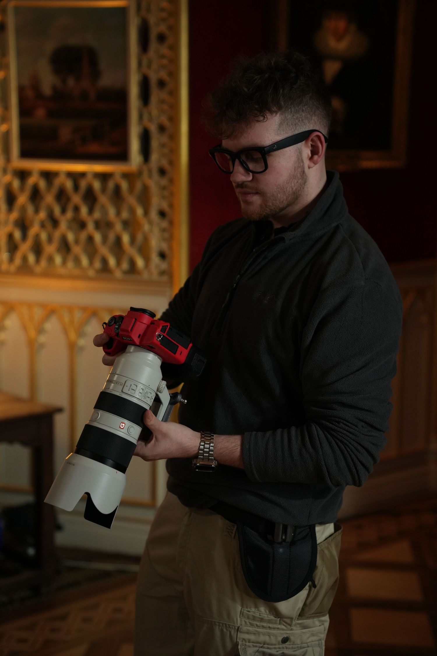 A young man with glasses examines a large professional camera lens in an ornate interior.