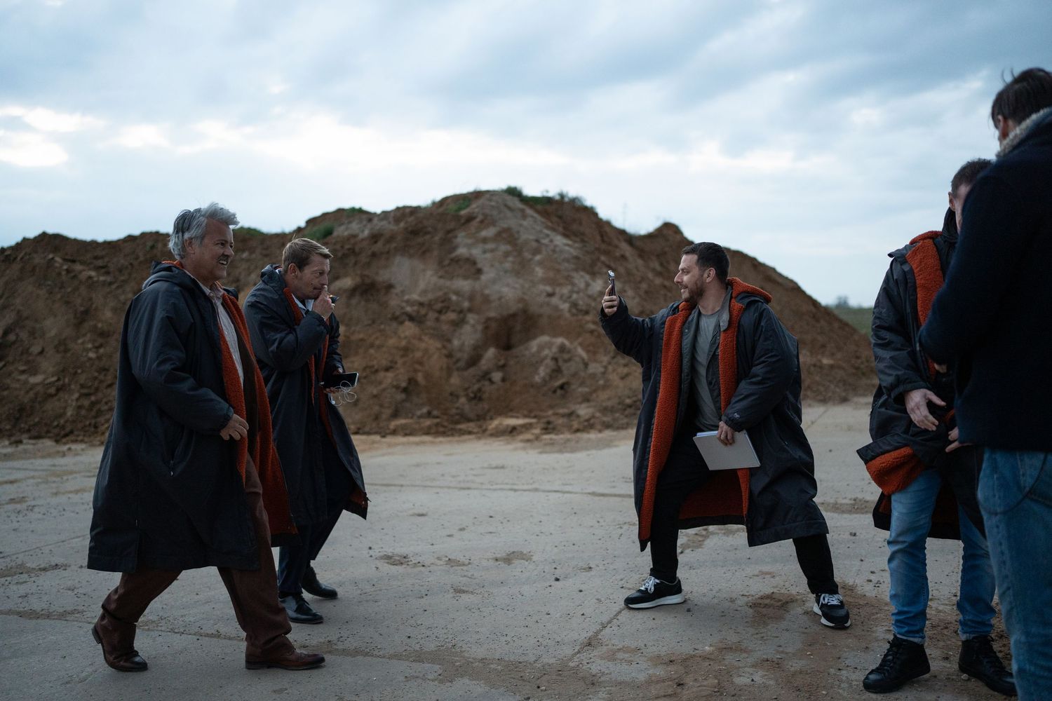 Film crew directs actors in matching dark coats at an outdoor quarry location shoot.