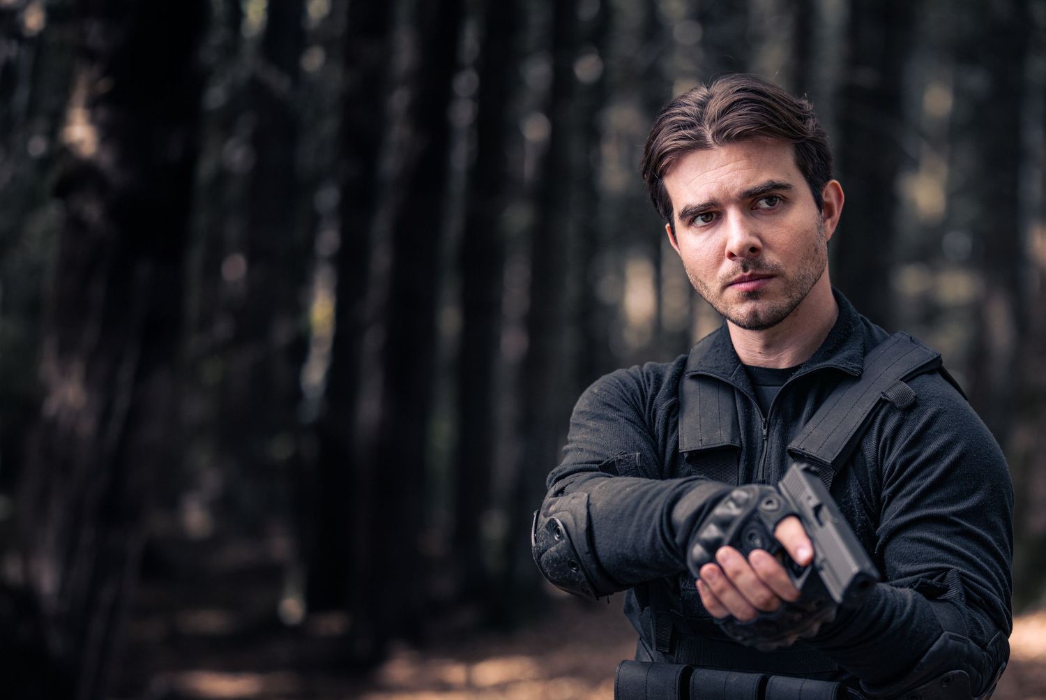 Man in tactical black jacket holding a pistol in a dark forest, intense action scene.