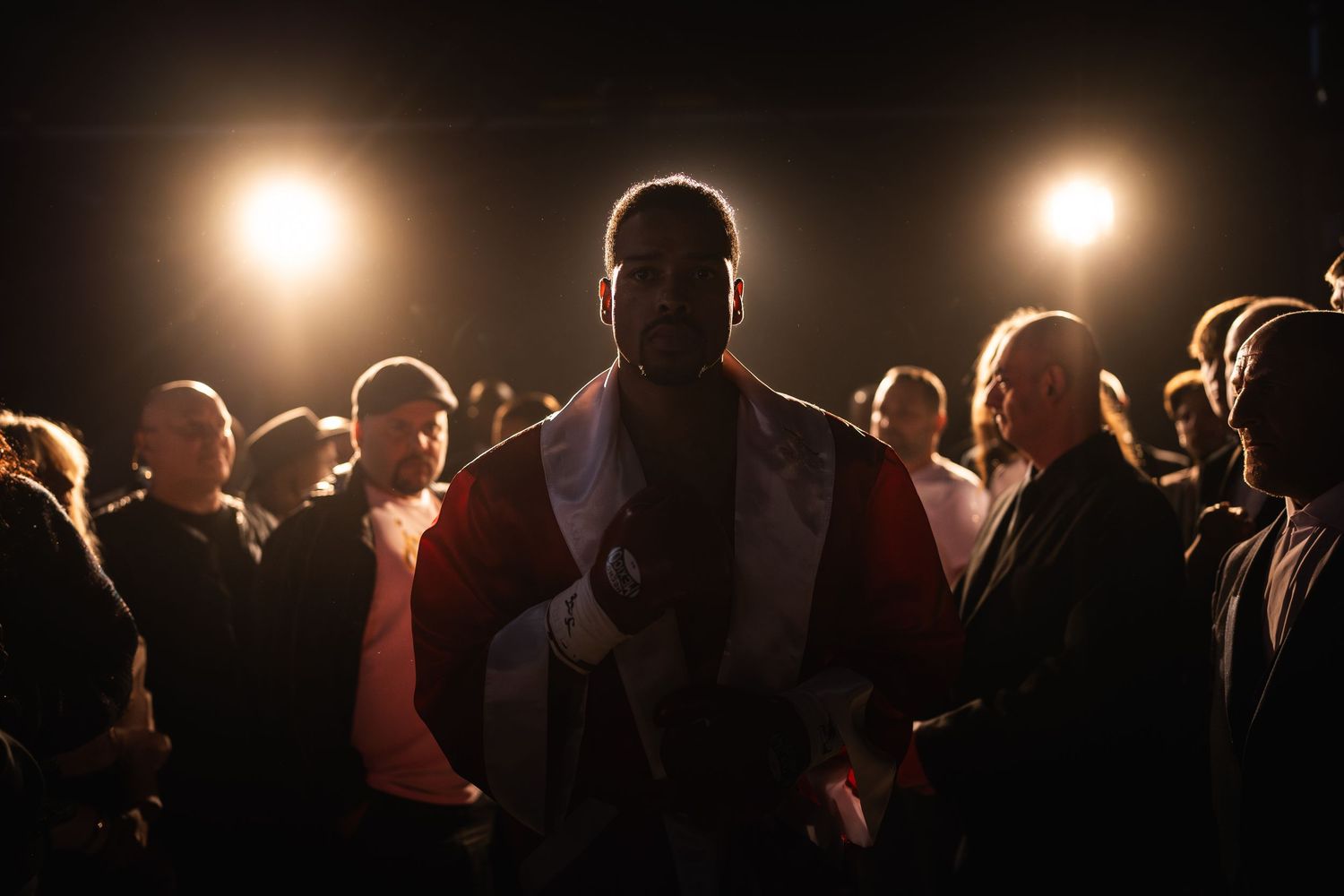 A boxer in a white and red robe stands center stage under dramatic spotlights surrounded by a crowd in a dark arena.
