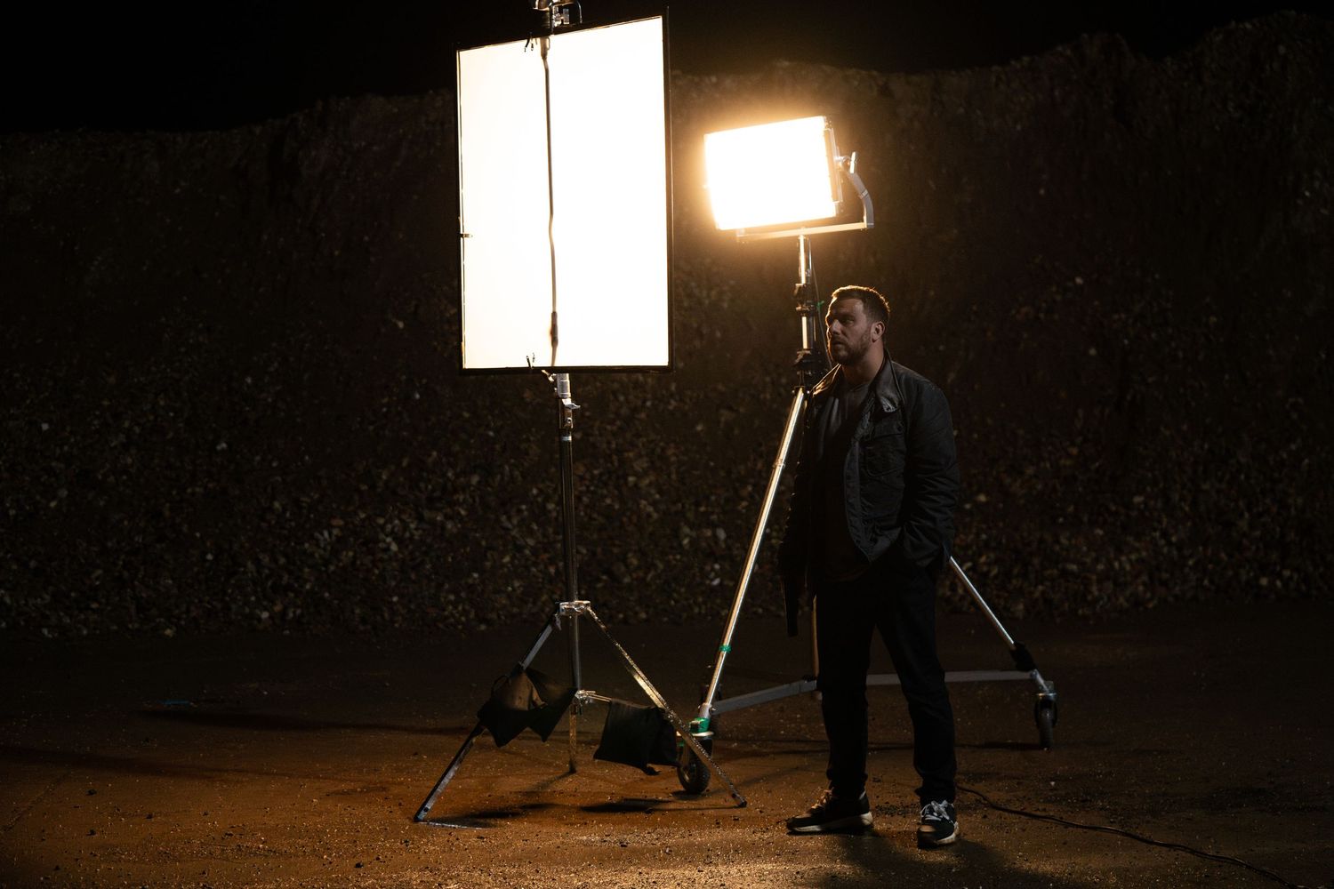 A filmmaker stands between two professional studio lights on tripods during a nighttime outdoor shoot.