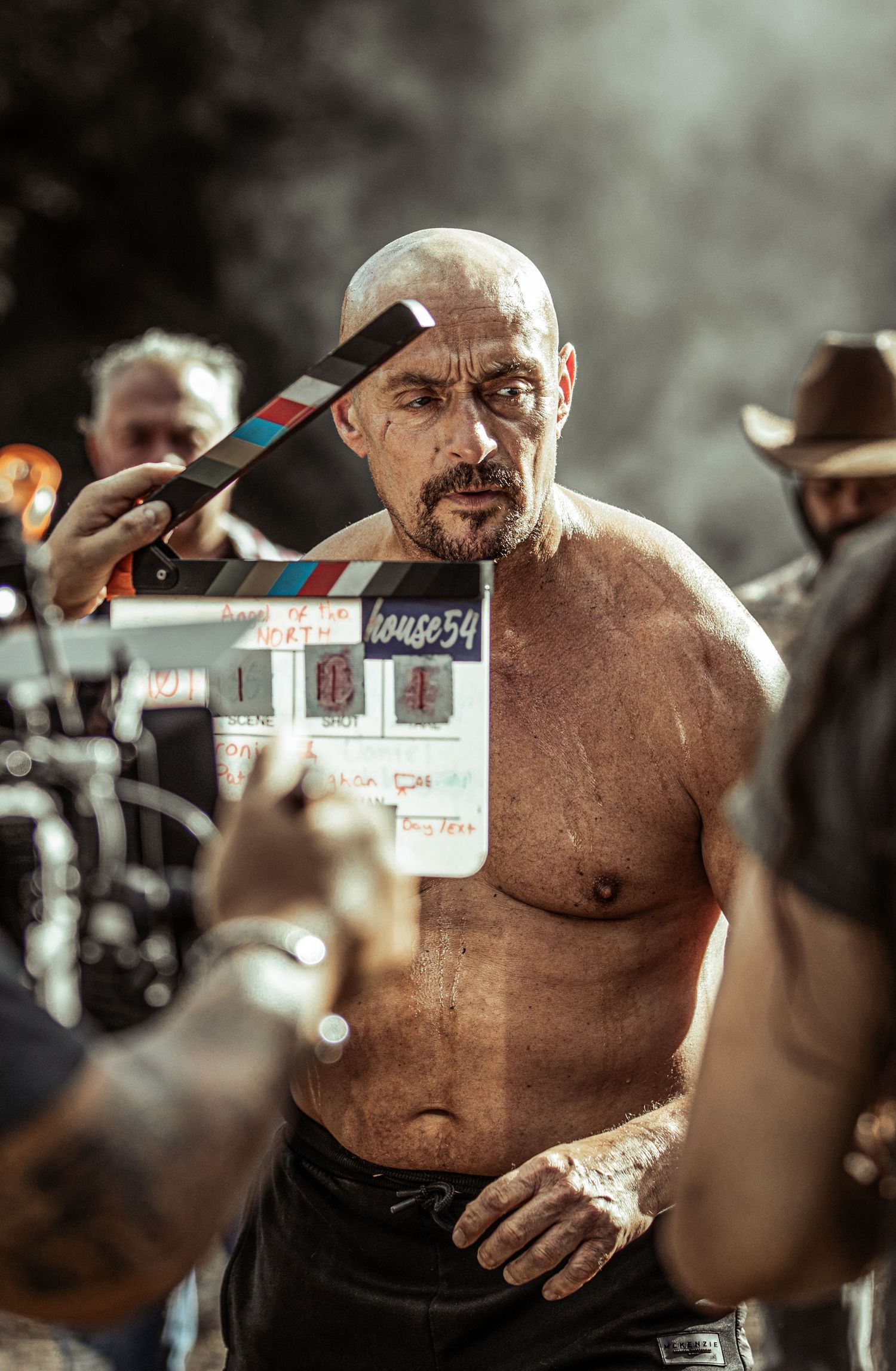 A bald shirtless man with tattoos stands on a film set as a clapperboard is held nearby.