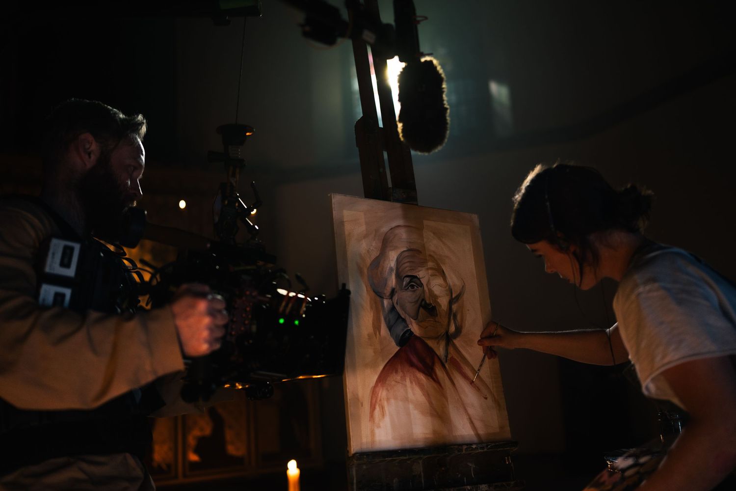 Cameraman films artist sketching a portrait by candlelight on a moody film set.