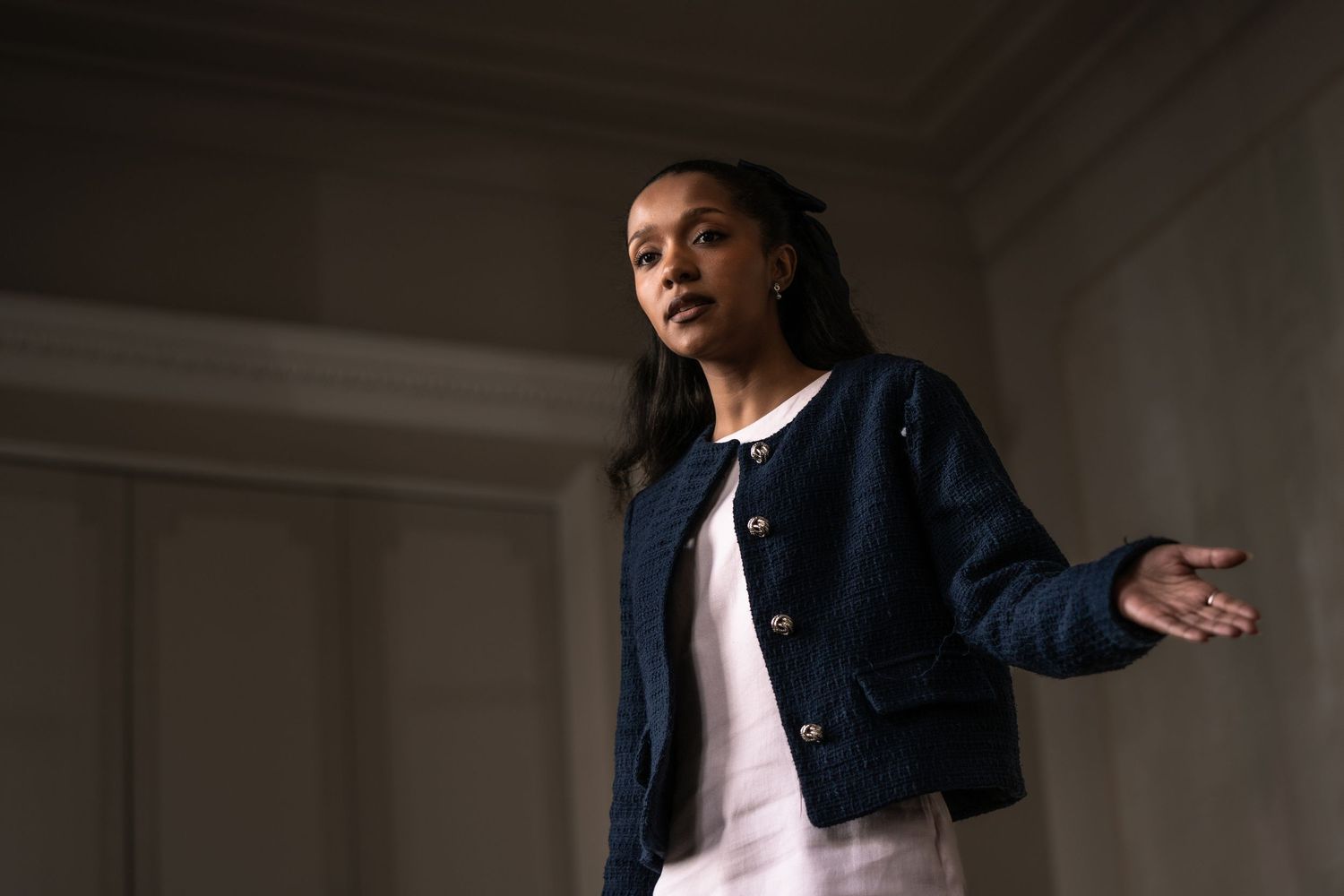 Young woman in navy blue jacket gestures with one hand in a dimly lit interior setting.