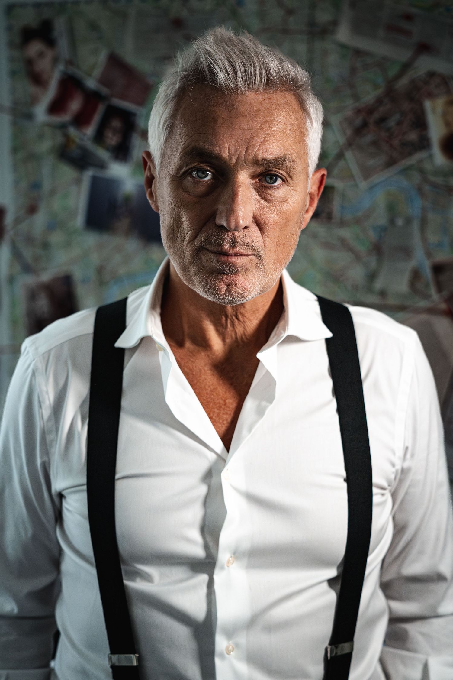 Silver-haired man in white shirt and suspenders stands before a detective investigation board.