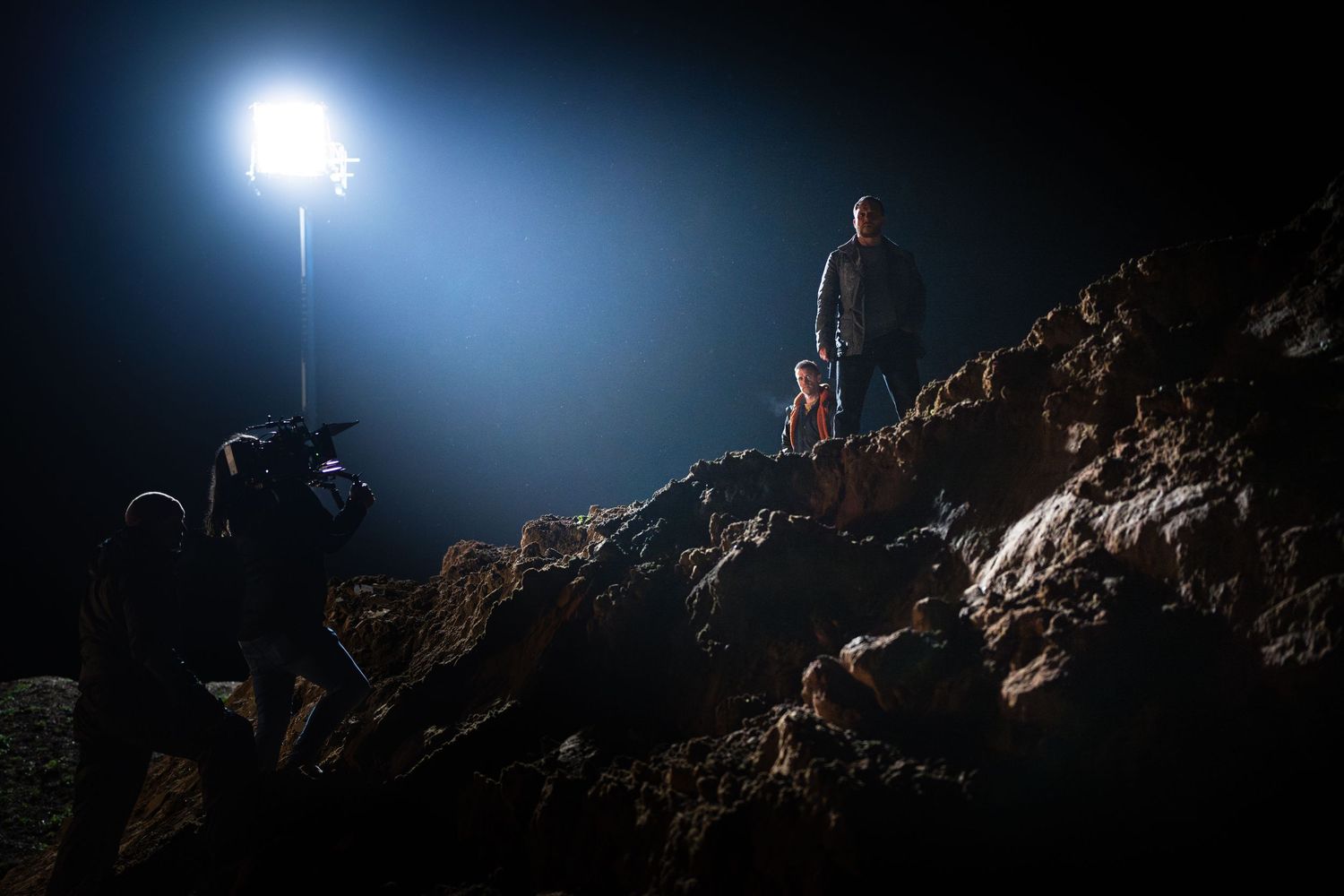 Film crew captures two actors standing on rocky terrain under a powerful spotlight at night.